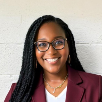 A woman with long braided hair, wearing glasses, a maroon blazer, and a white top, smiles in front of a light-colored wall.