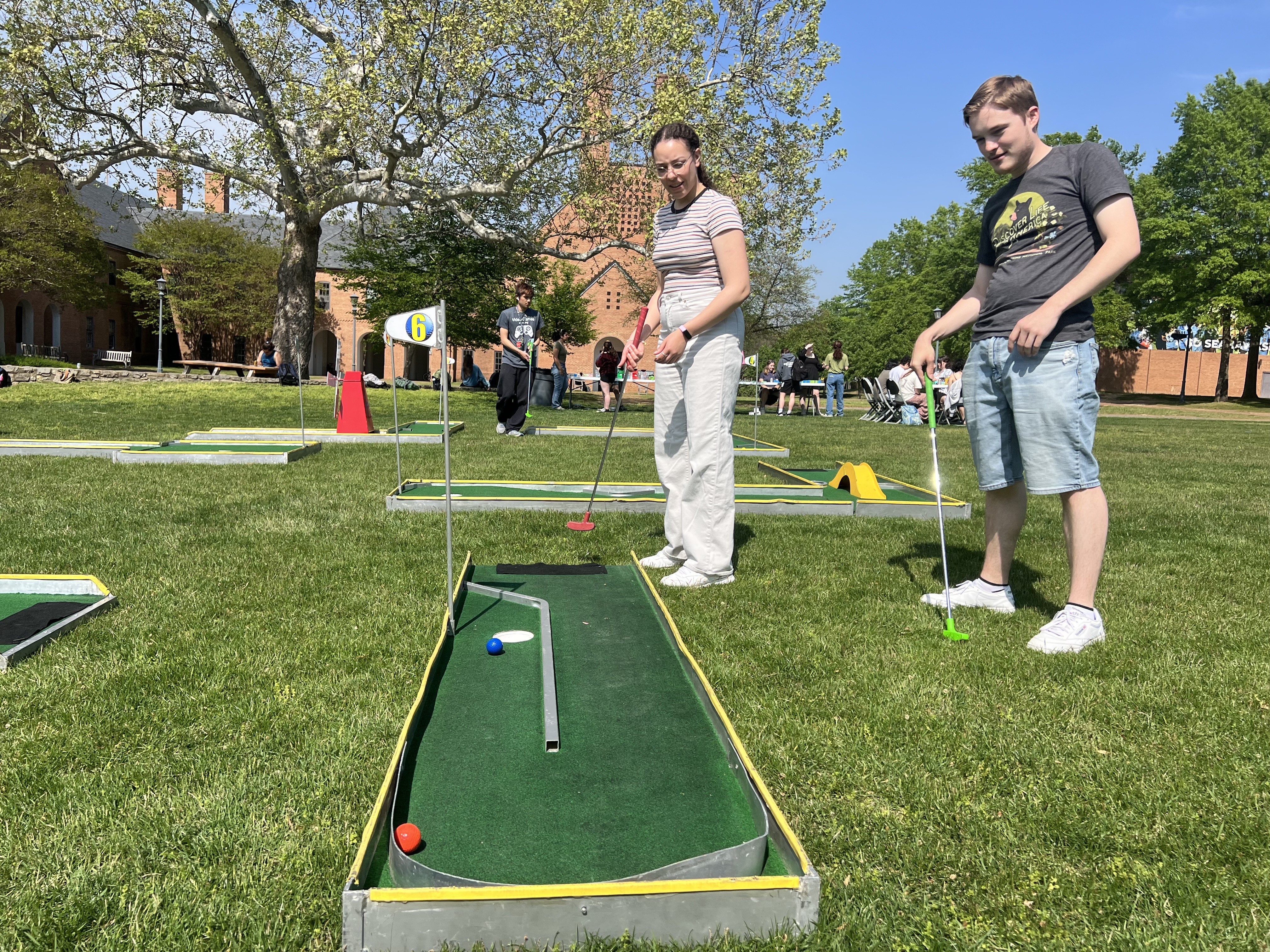 Two people play mini-golf on a portable course set up on grass, with trees and buildings in the background on a sunny day.