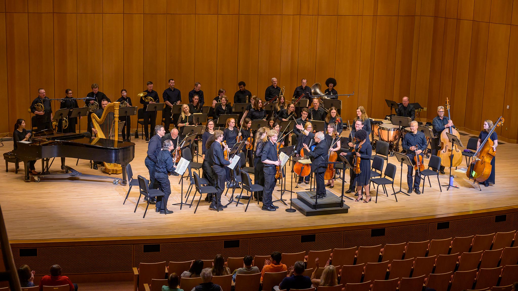 An orchestra performs on stage in a concert hall, with musicians playing various instruments including strings, woodwinds, brass, harp, and piano. Audience members are seated in front.