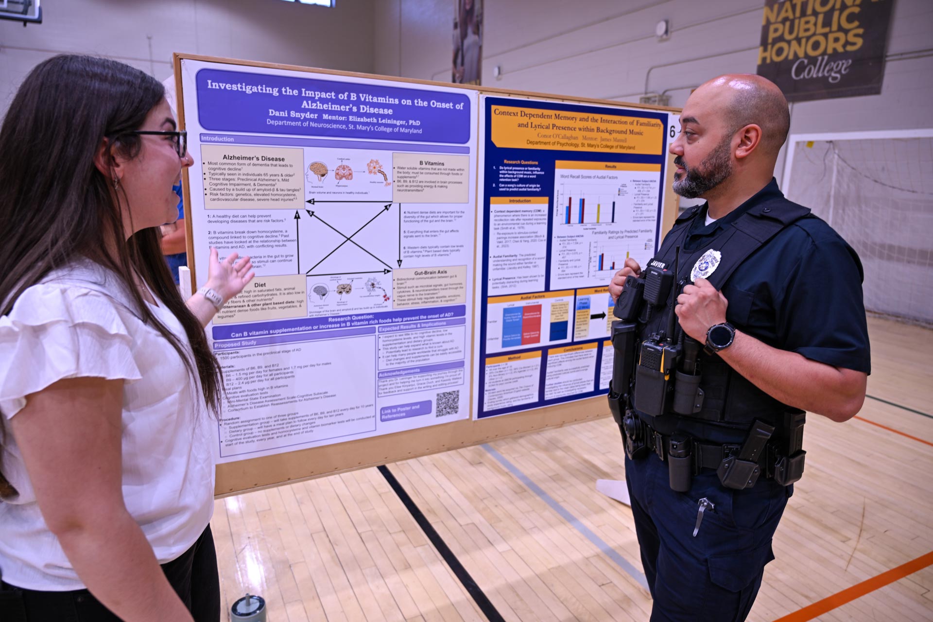A woman explains a scientific research poster to a uniformed police officer in a gymnasium with a volleyball net in the background.