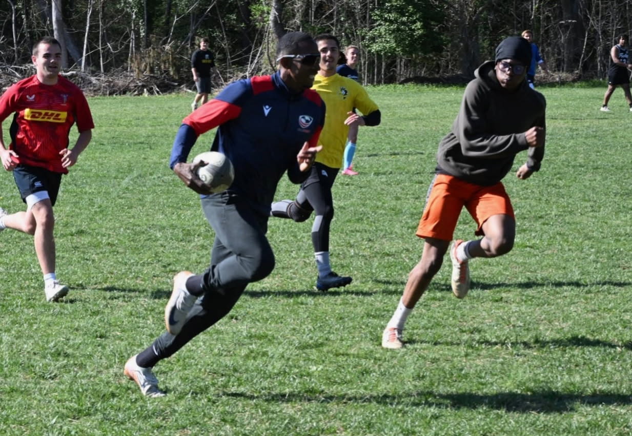 A group of men playing rugby on a grassy field. One player in a dark shirt is running with the ball, pursued by others. Trees and additional players are visible in the background.