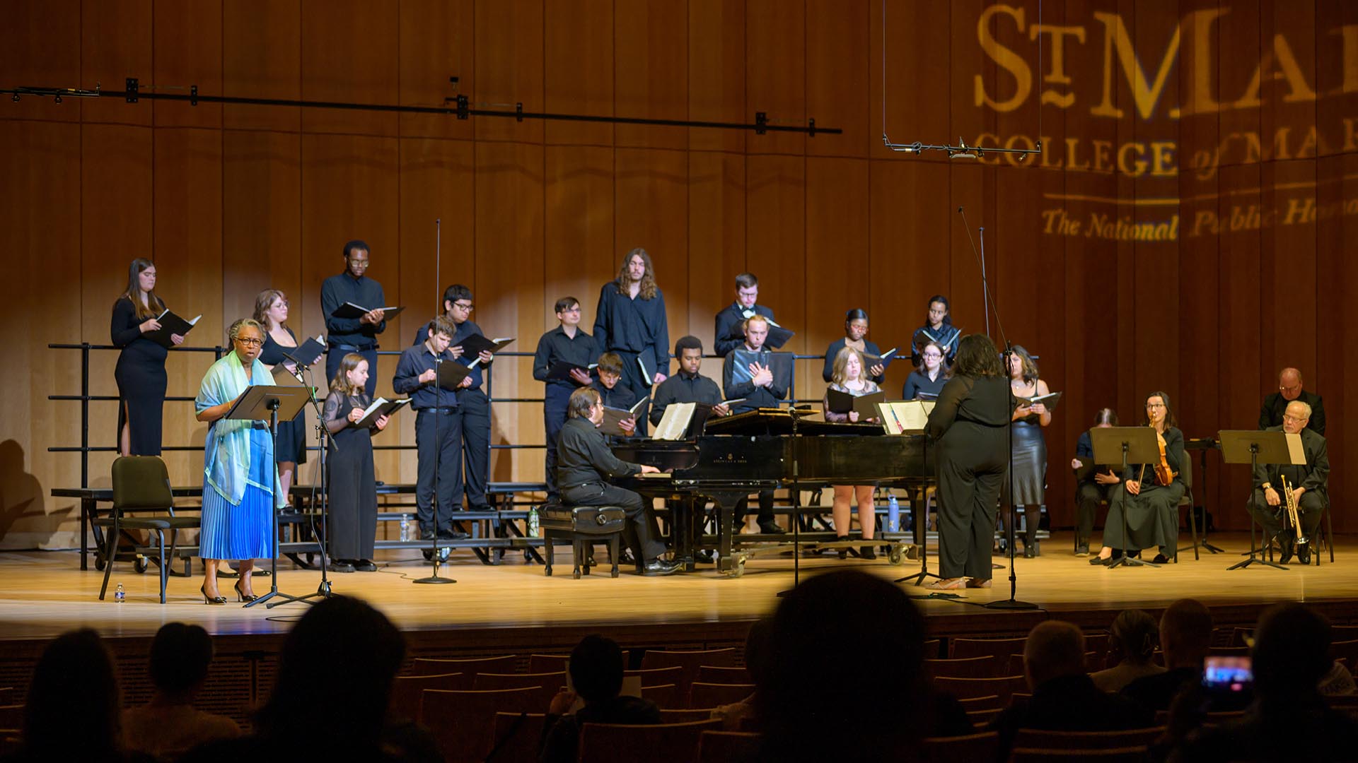 A choir and pianist perform on stage in a concert hall, with a conductor leading and an audience seated in the foreground.