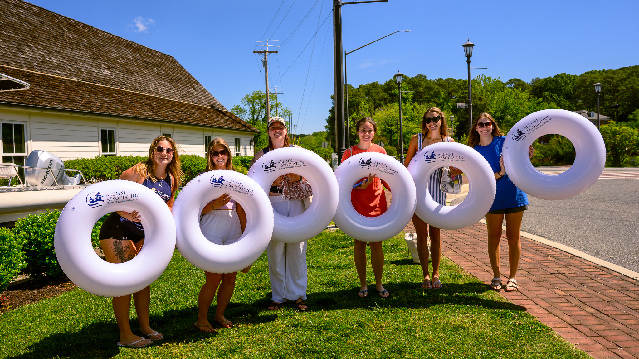Six people standing on grass, each holding a large white inflatable tube with a logo, in front of a building and street on a sunny day.