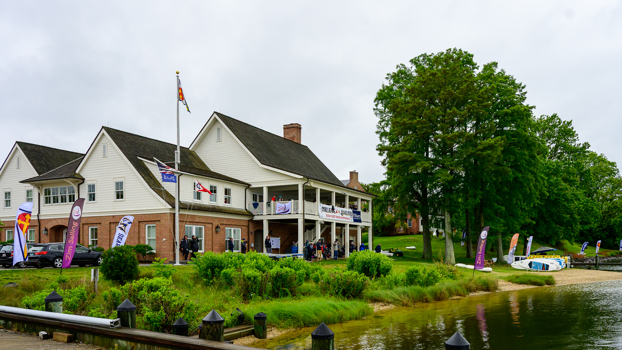 A two-story brick and white building with flags and banners hosts an event near a sandy waterfront, surrounded by greenery and trees.