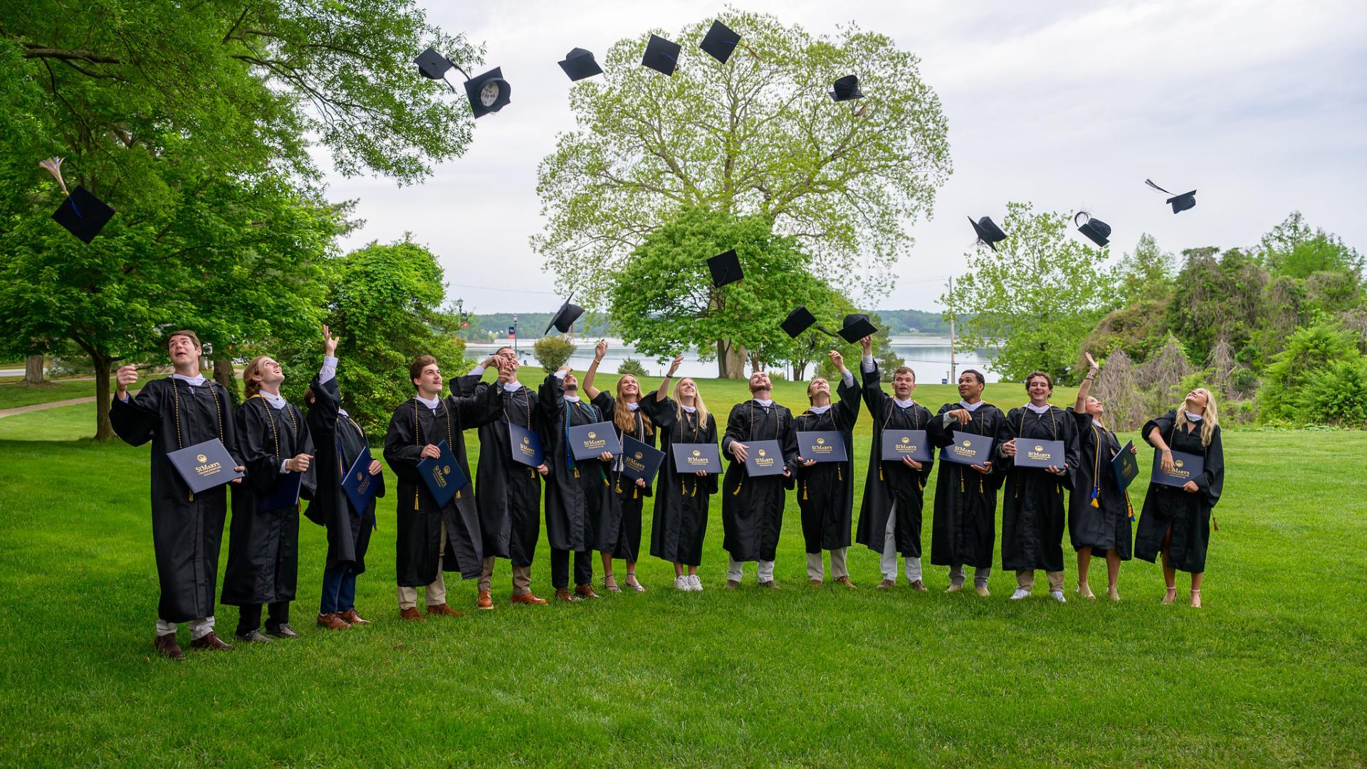A group of graduates in caps and gowns stands on grass, throwing their caps in the air while holding diplomas, with trees and a lake in the background.