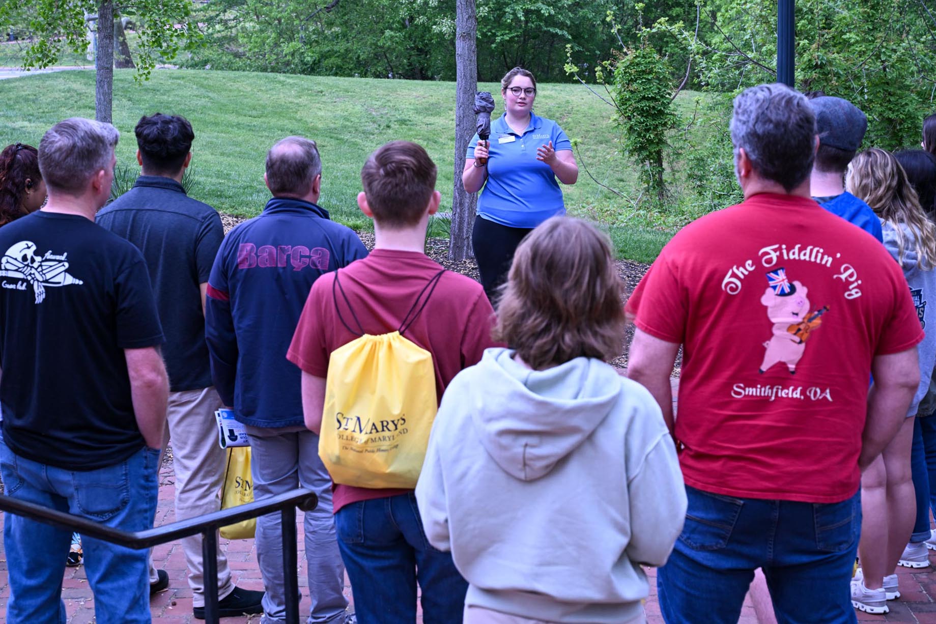 A tour guide speaks to a group of people outside on a brick walkway, with grass and trees visible in the background.