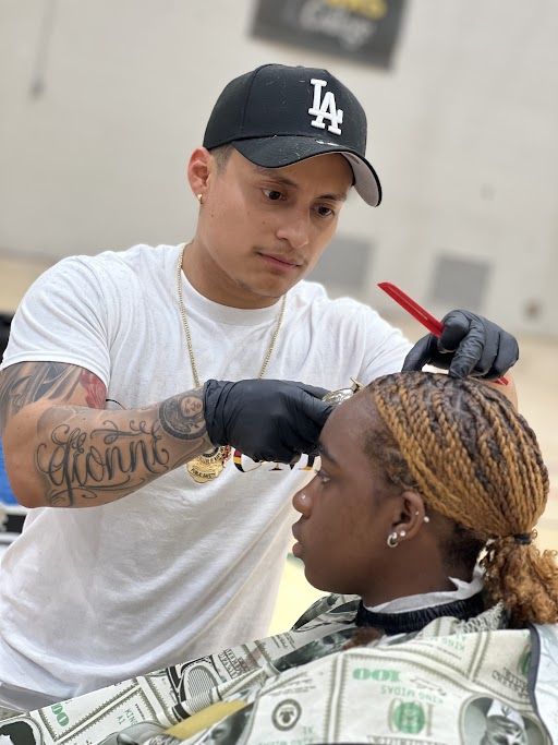 A barber wearing gloves and a black LA cap trims a seated client's braided hair with scissors and a comb.