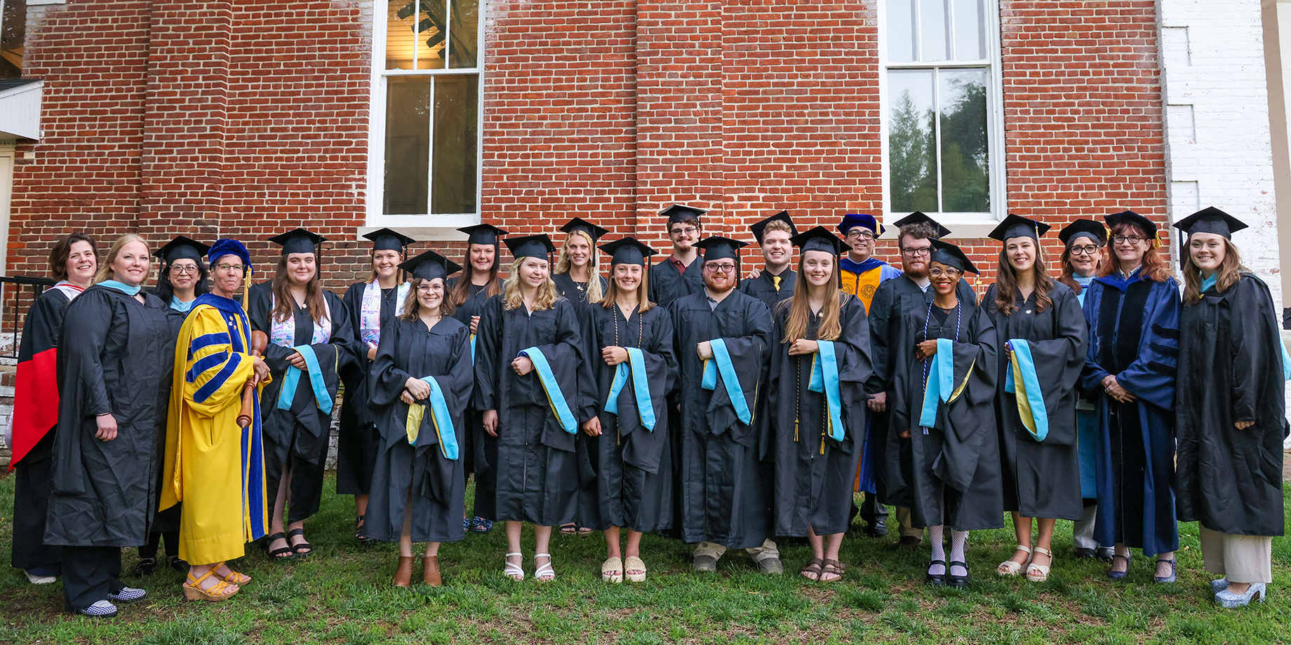 A group of graduates and faculty members in academic regalia pose for a photo in front of a brick building.