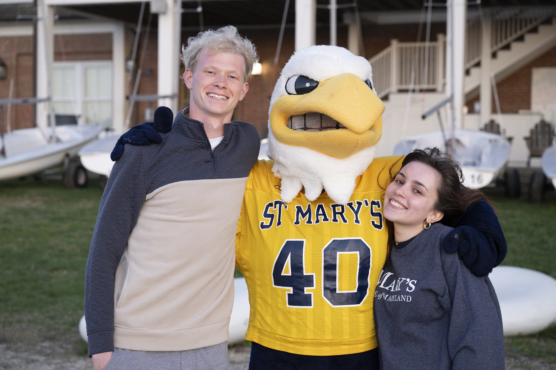 Two students stand smiling next to a person in an eagle mascot costume wearing a St. Mary's jersey numbered 40, with boats and a building in the background.