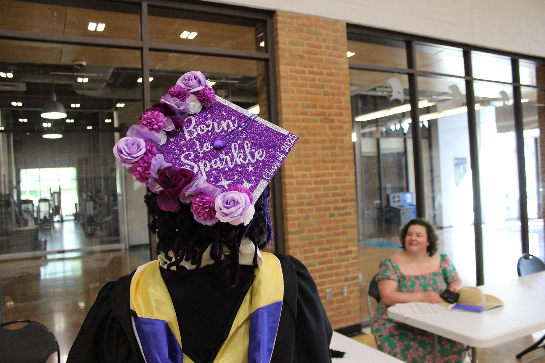 A graduate wearing a decorated cap reading "Born to Sparkle" stands near a table where a woman is seated, inside a building with glass walls and brick accents.