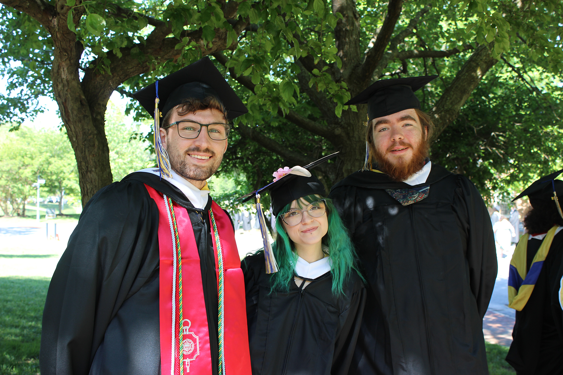 Three graduates in caps and gowns stand outdoors under a tree, smiling at the camera on a sunny day.