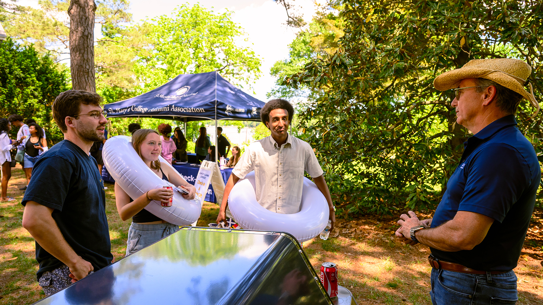 Four people stand outdoors by a booth and canopy tent; two hold inflatable swim rings, and one man wears a straw hat. Trees and greenery surround the area.