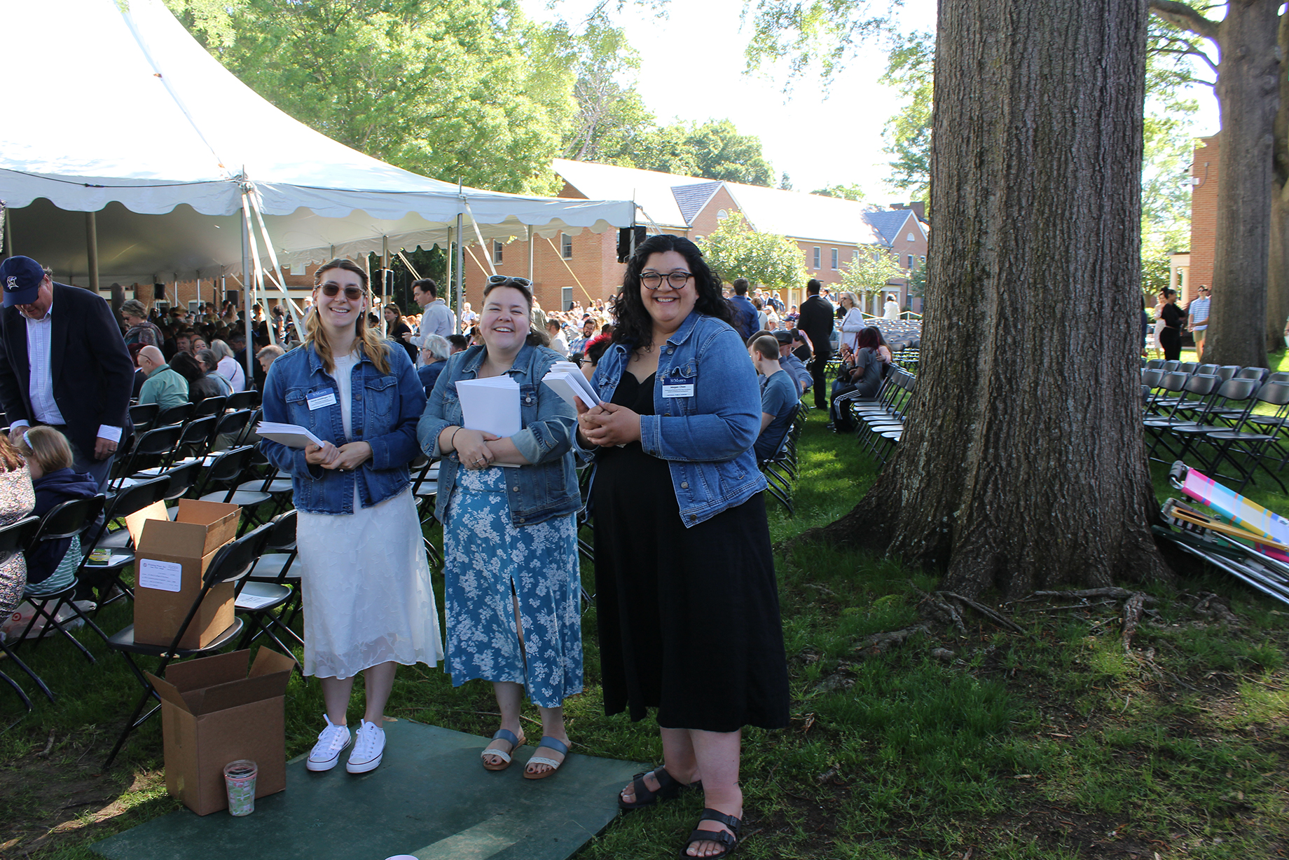Three women in denim jackets stand and smile near a tree at an outdoor event with chairs and a tent in the background.