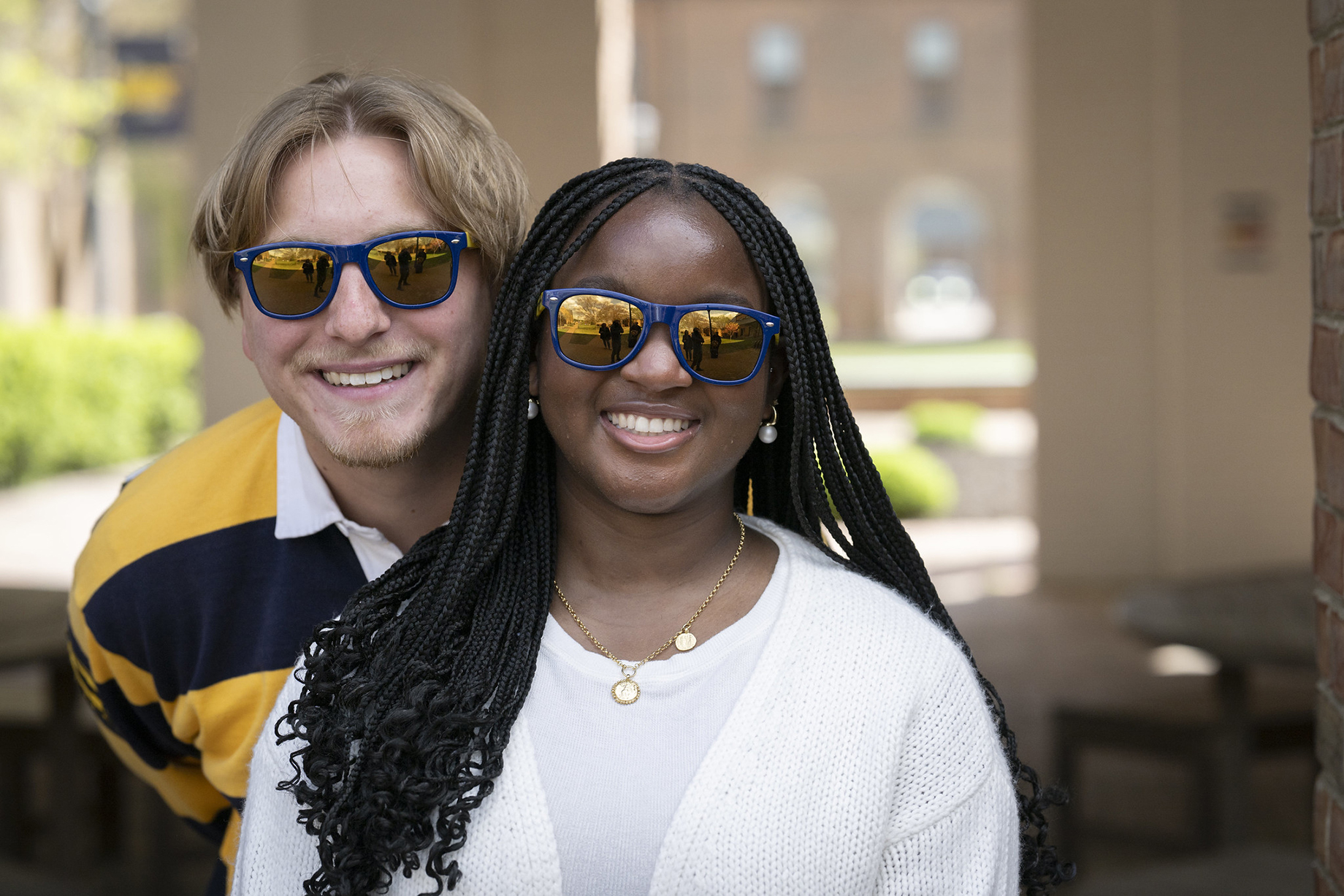 Two people wearing sunglasses are smiling outdoors. The person in front has long braids and white clothing; the person behind has light hair and a striped shirt.