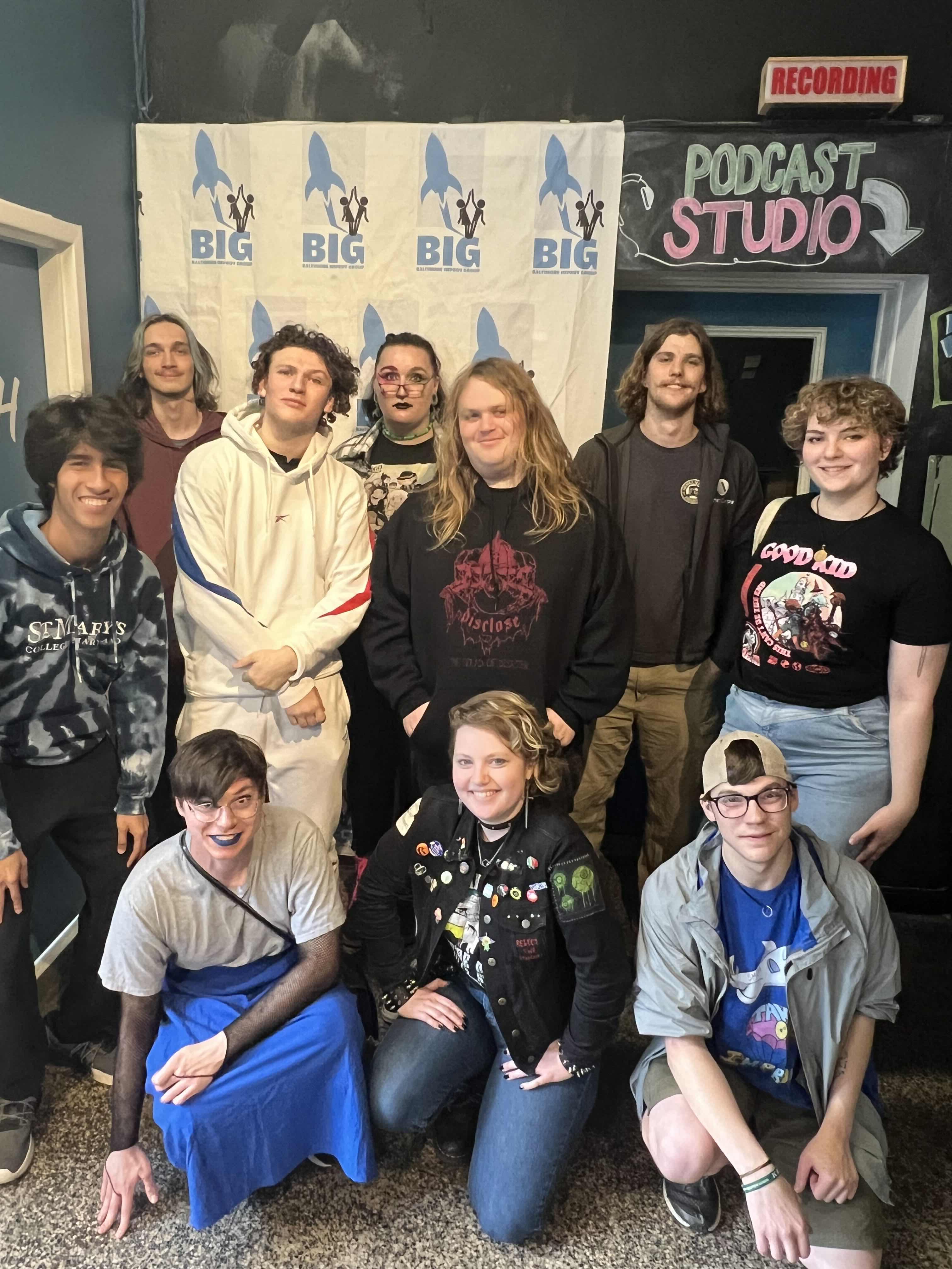 A group of eleven young adults pose indoors in front of a "BIG" banner and a "Podcast Studio" sign.