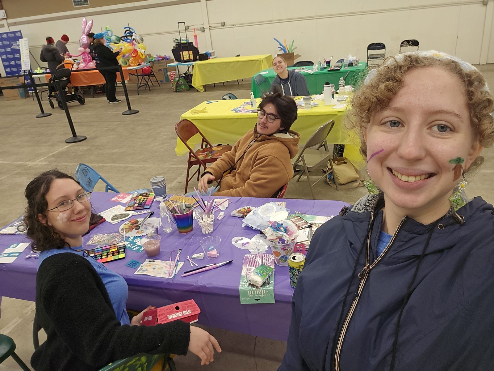 Four people sit and smile at a table covered with art supplies and snacks in a large indoor space decorated with colorful tables and balloons.
