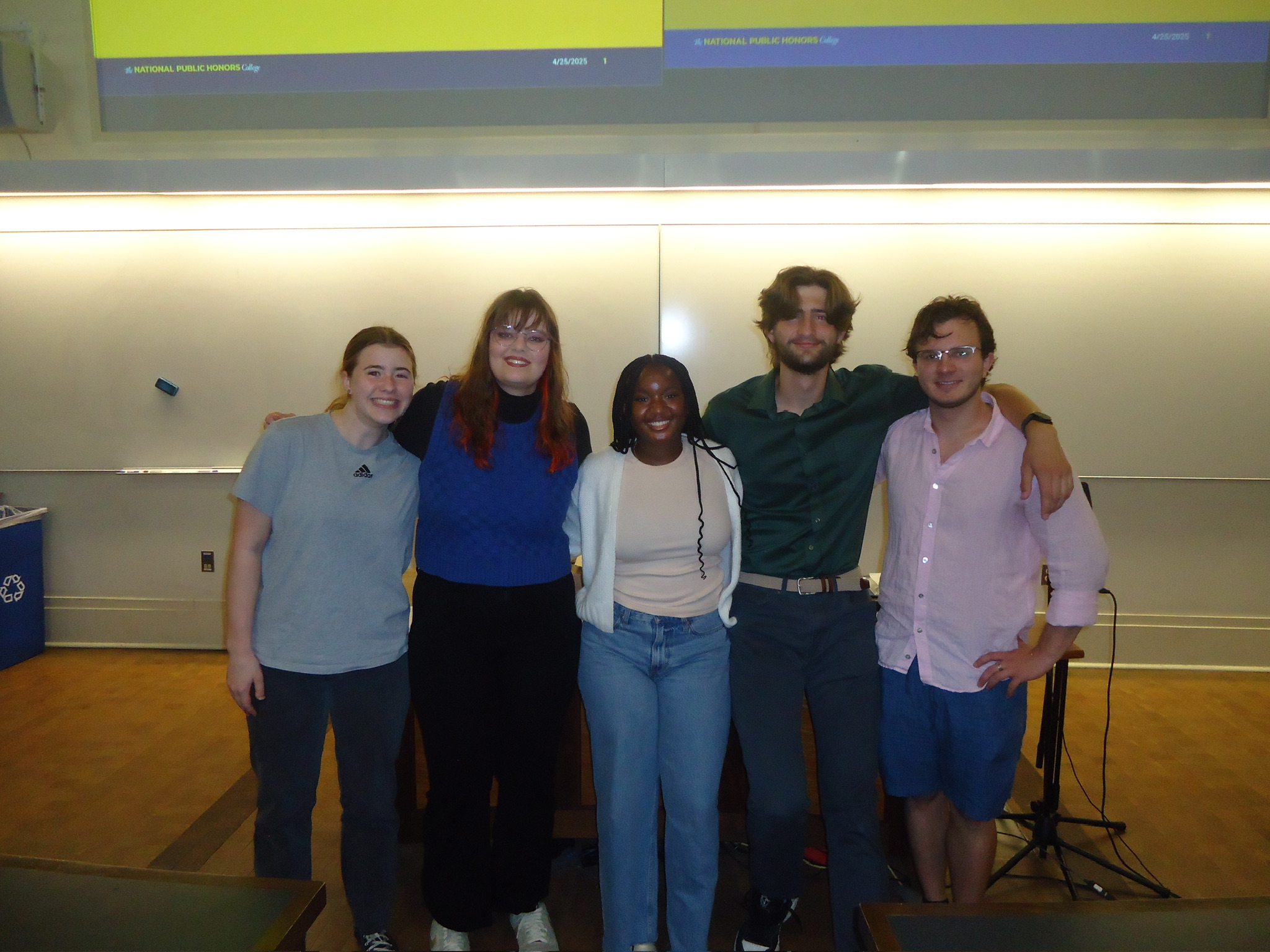 Five people stand side by side in a classroom, smiling at the camera, with a whiteboard and projection screens in the background.