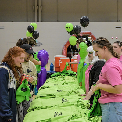 People distribute green T-shirts and bags at a table decorated with alien-themed balloons and inflatables during an indoor event.