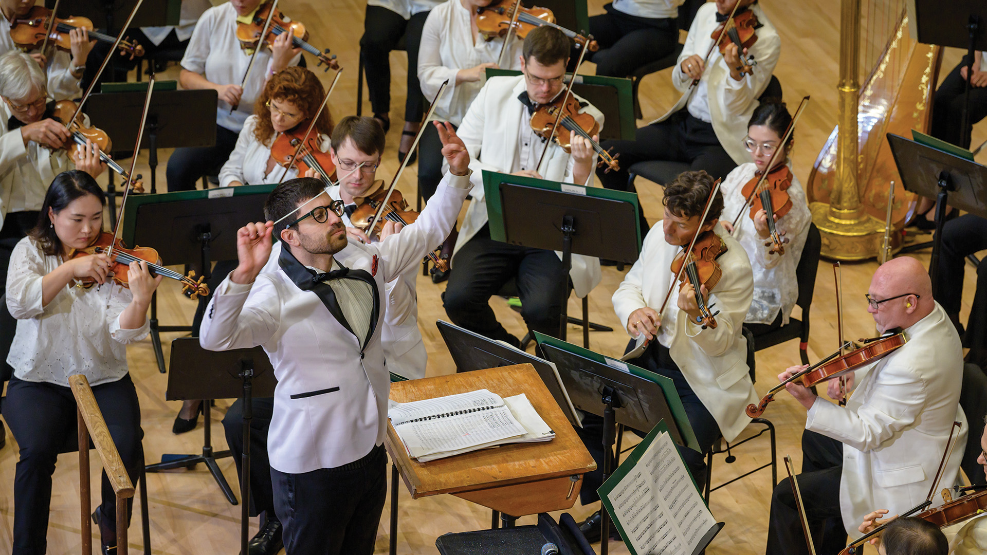 A conductor leads an orchestra of musicians playing string instruments. Most musicians are wearing white jackets and are focused on their sheet music.