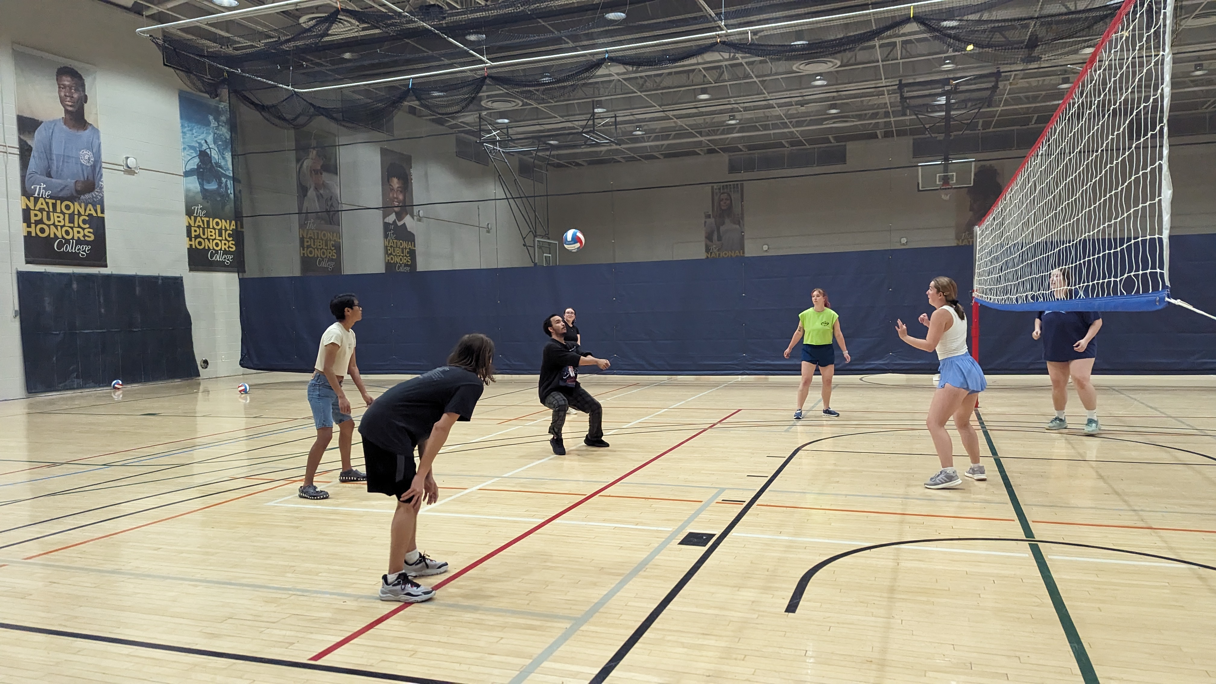 A group of six people play volleyball indoors on a gym court, with one player preparing to hit the ball over the net.