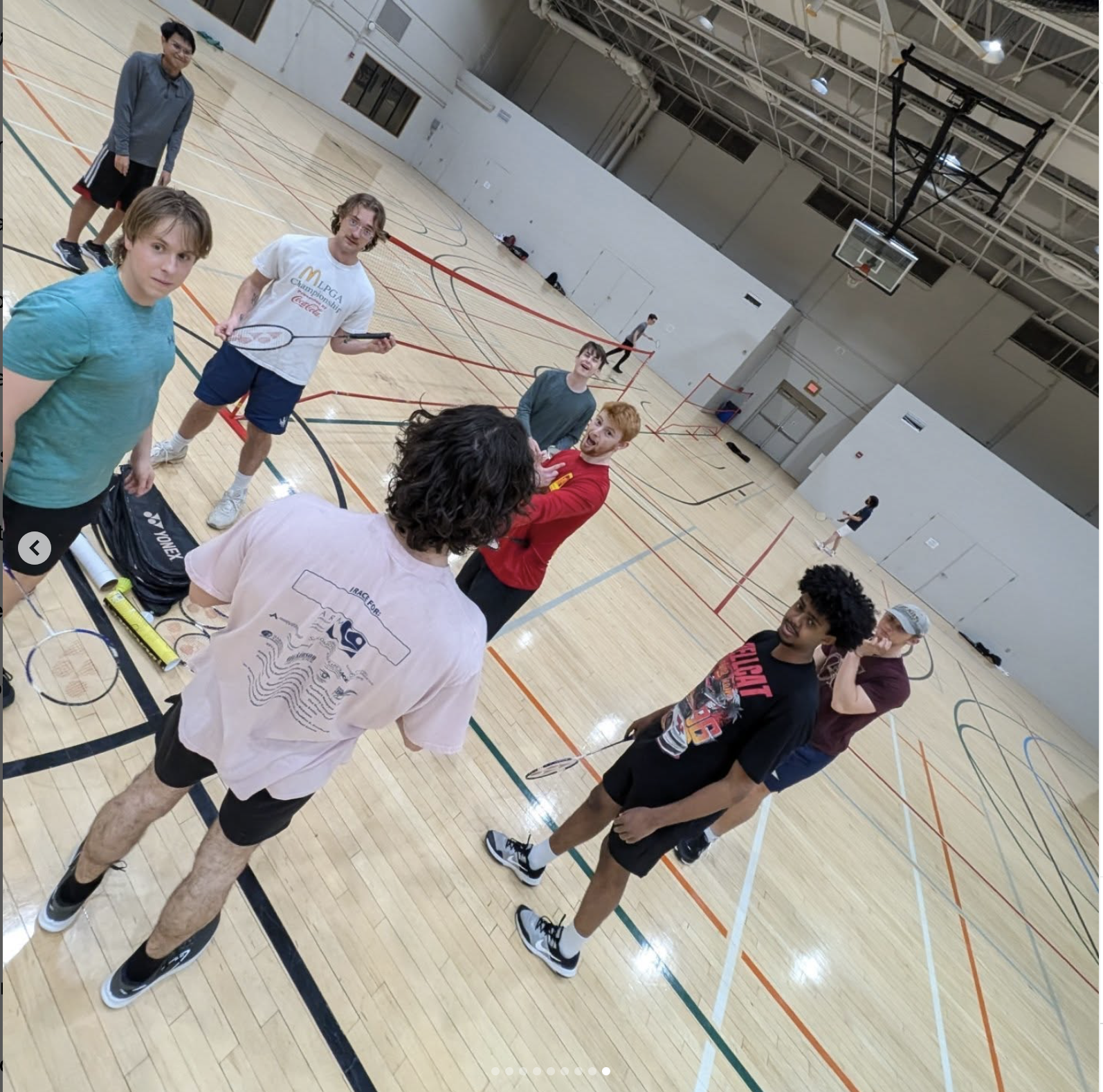 A group of eight people stand in a gymnasium holding badminton rackets, with bags and shuttlecocks on the floor. Indoor courts and equipment are visible in the background.