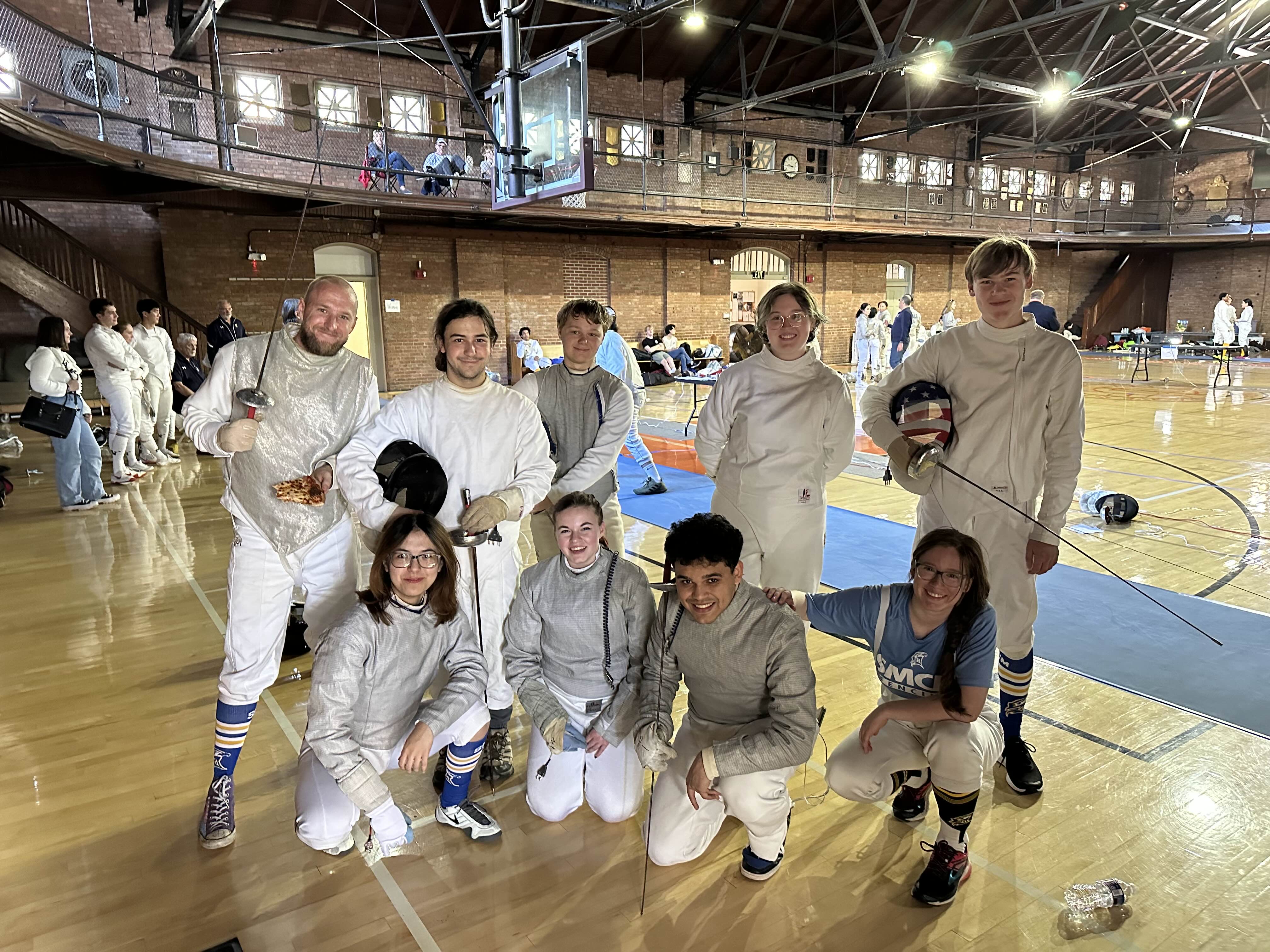A group of fencers in full gear pose together on a gym floor, with fencing equipment and spectators visible in the background.