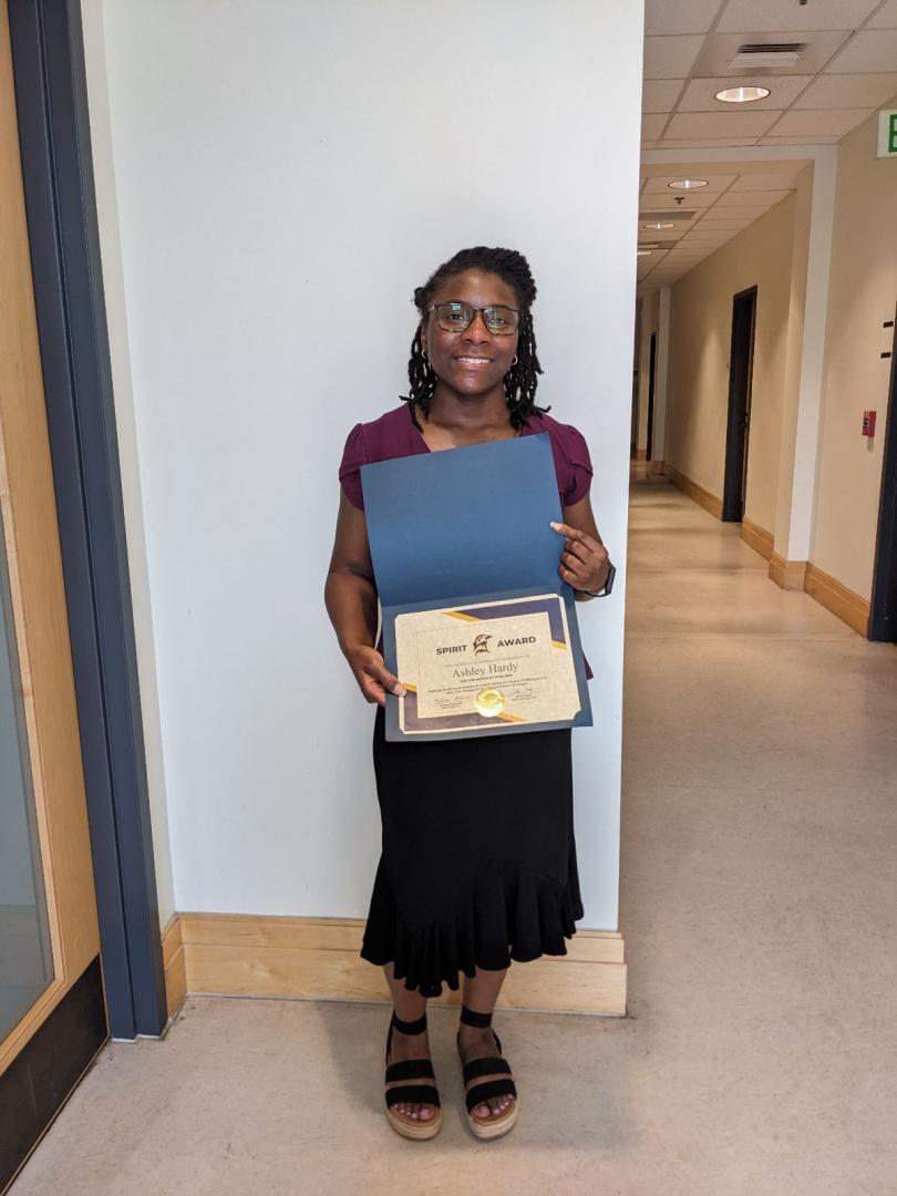 A person stands in a hallway holding an open folder with a "Spirit Award" certificate, smiling at the camera.