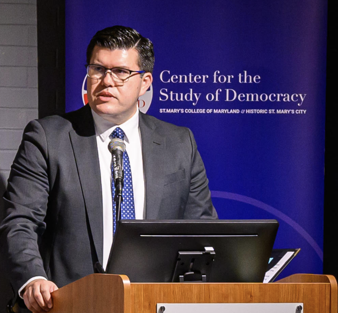 A man in a suit speaks at a podium with a microphone in front of a sign for the Center for the Study of Democracy at St. Mary's College of Maryland.