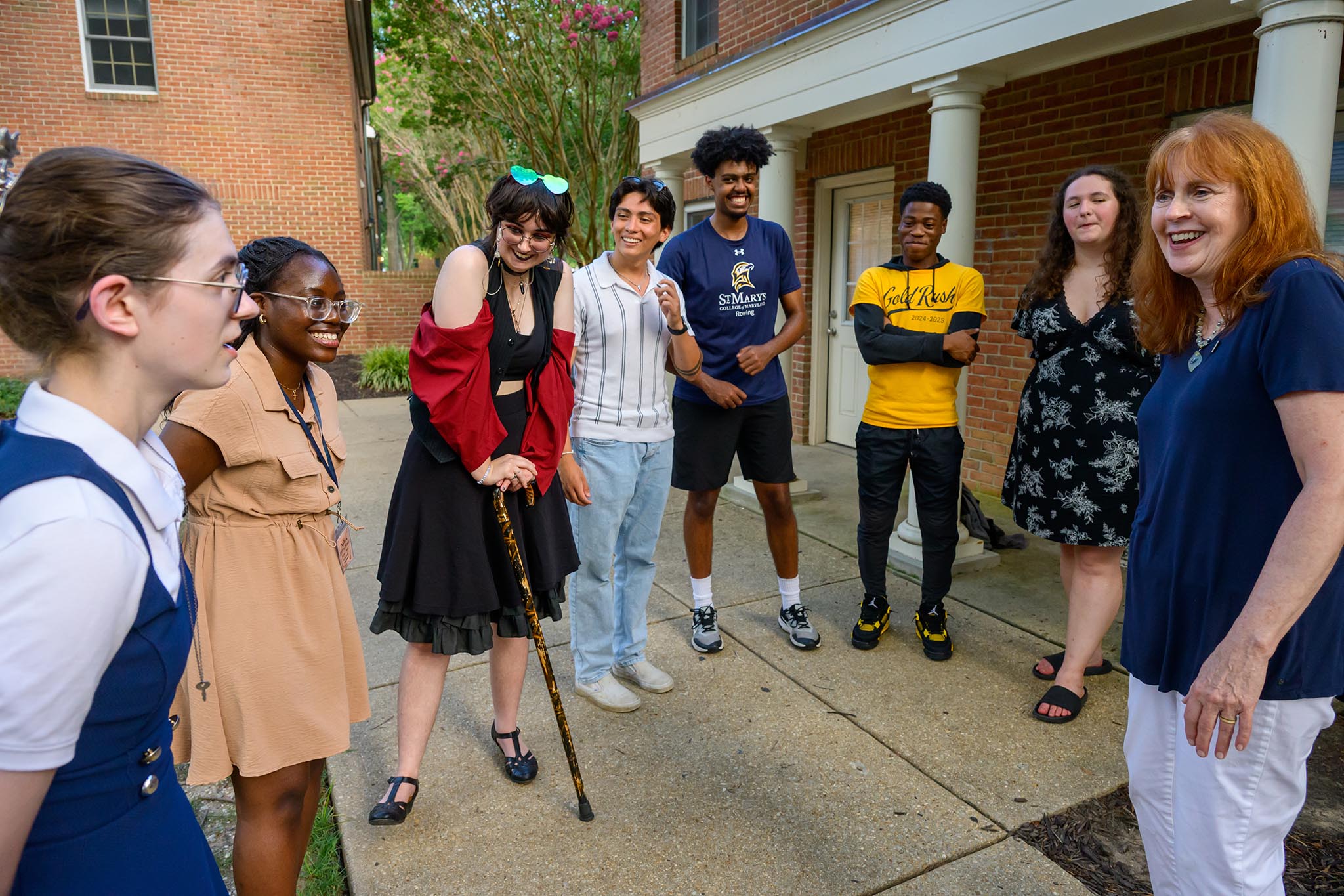 A group of seven young people and one adult woman stand outside a brick building, talking and smiling together.