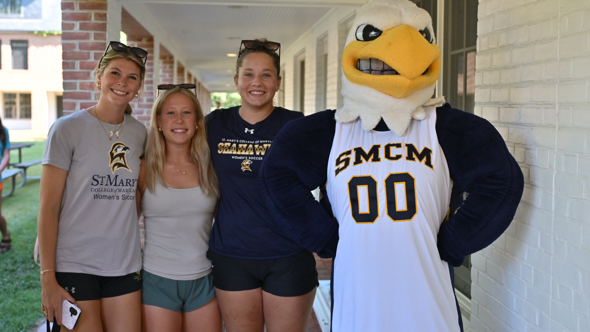 Three young women stand next to a person in a Seahawk mascot costume wearing an "SMCM 00" jersey, posing for a photo outdoors by a brick building.