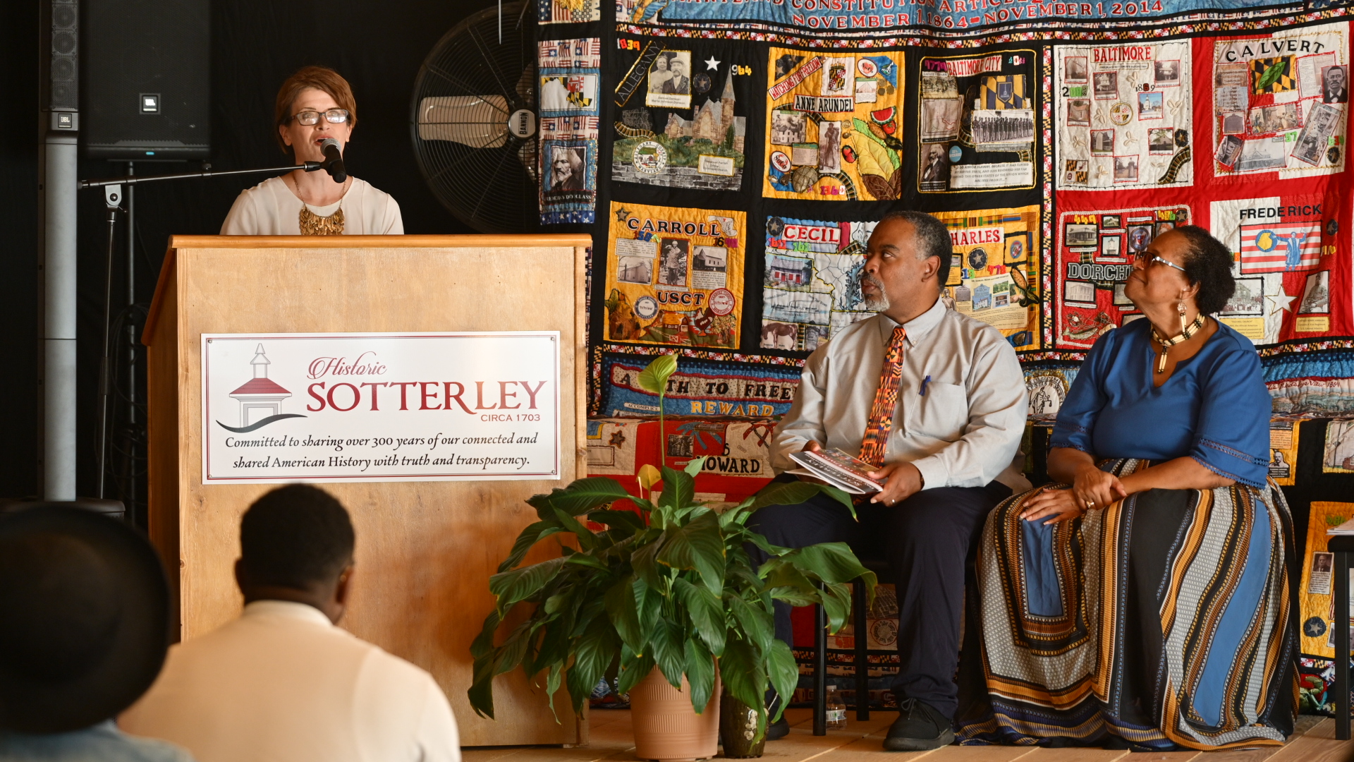 A woman speaks at a podium labeled “Historic Sotterley” while a man and woman sit onstage in front of a colorful quilt backdrop.