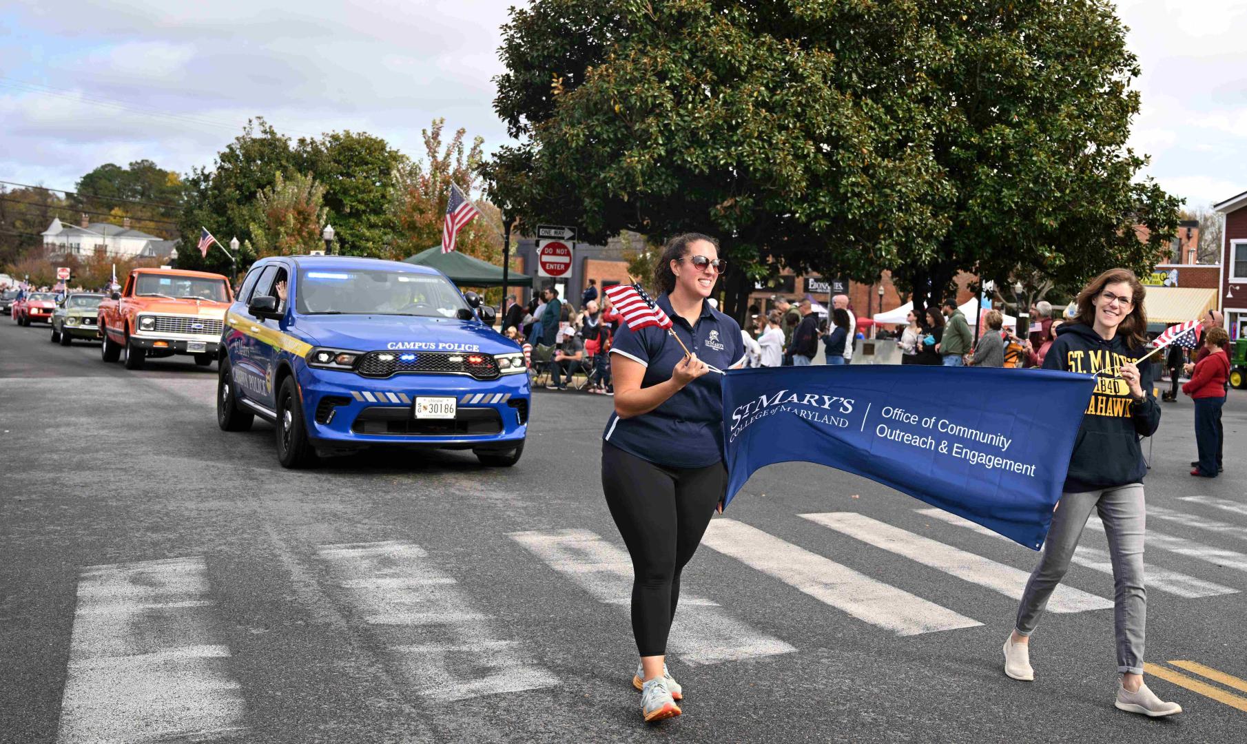 People walk in a parade, holding a “St. Mary’s College of Maryland” banner, with a police vehicle and participants carrying American flags in the background.