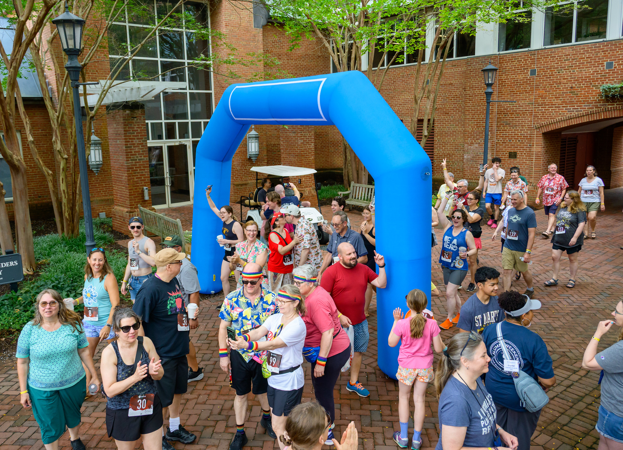 A group of people gathers under and around a blue inflatable arch outdoors, some wearing race bibs, casual clothes, and holding drinks, with a mix of cheering and smiling participants.
