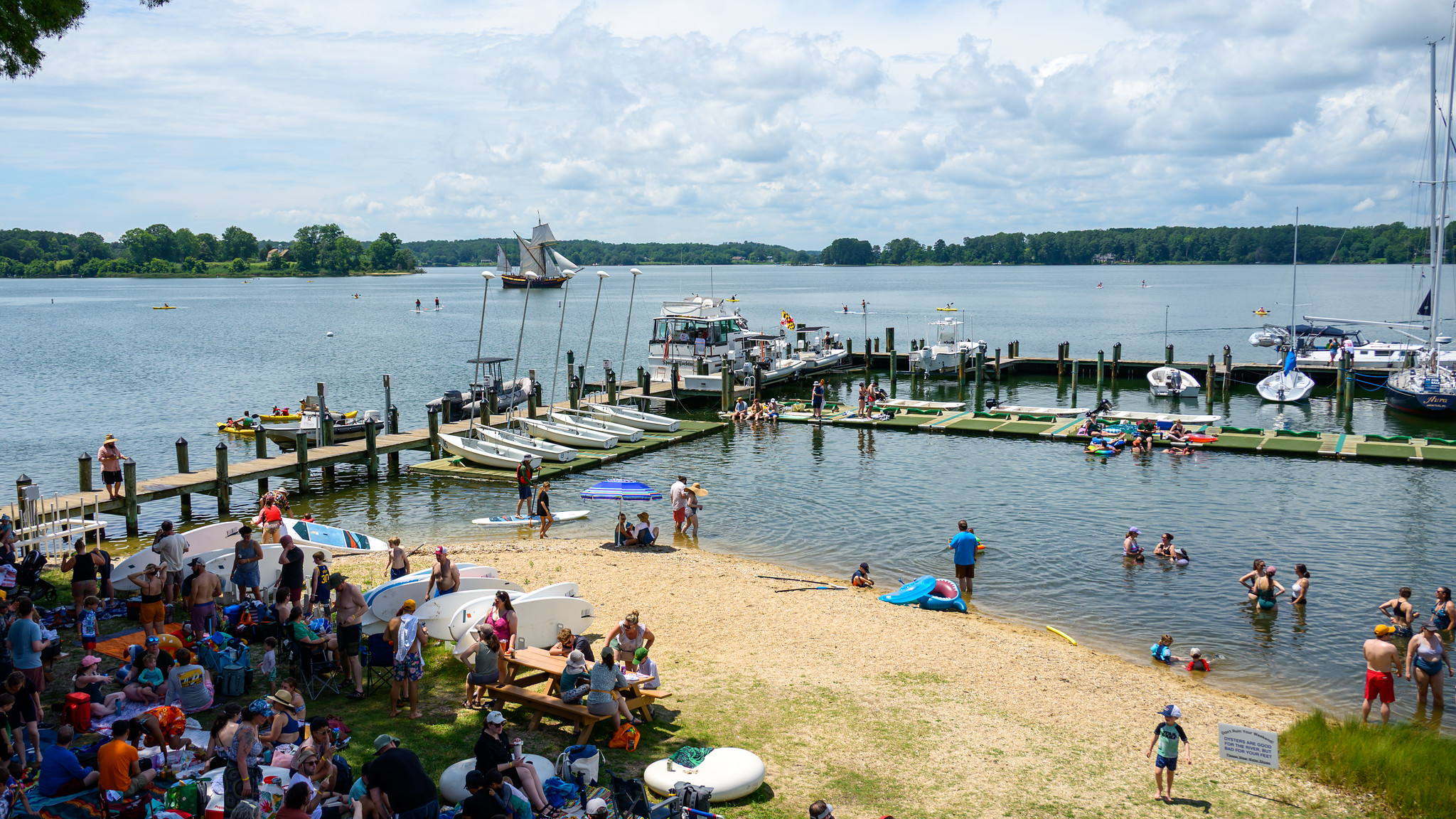 Crowd of people at a lakeside beach and marina, with boats docked, people swimming, and groups gathered on the sand and grass under a partly cloudy sky. A 17th-century replica sailing ship sails in the background.