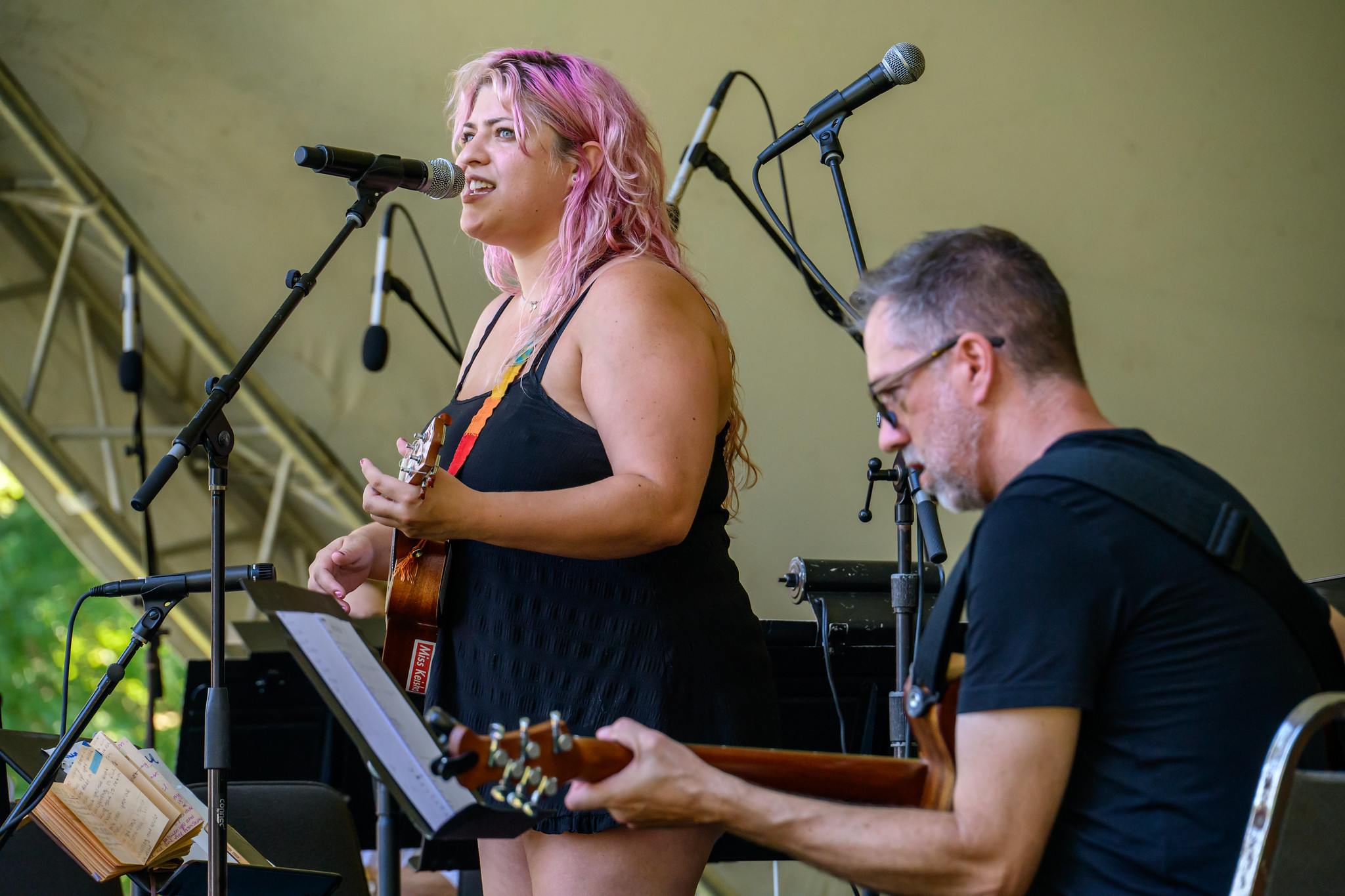 A woman with pink hair sings and plays ukulele on stage next to a man playing guitar. Microphones and music stands are visible in the background.