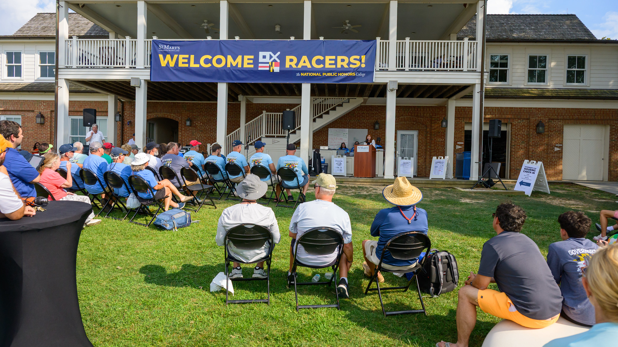 A group of people sit on chairs outdoors facing a stage where a speaker addresses them under a large banner that reads "WELCOME RACERS!"