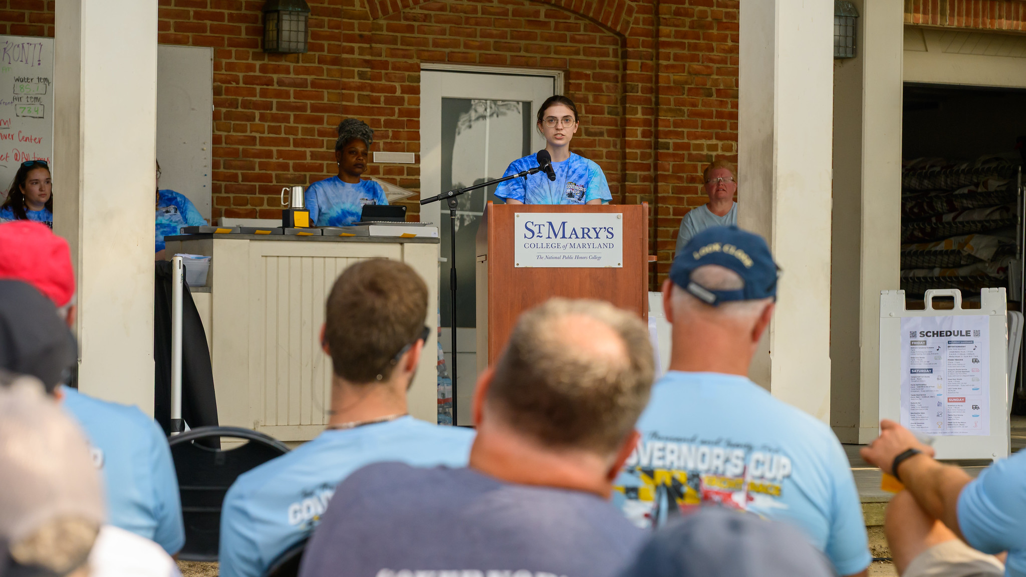 A person speaks at a podium labeled "St. Mary's College of Maryland" during an outdoor event, with people seated and listening in the foreground.