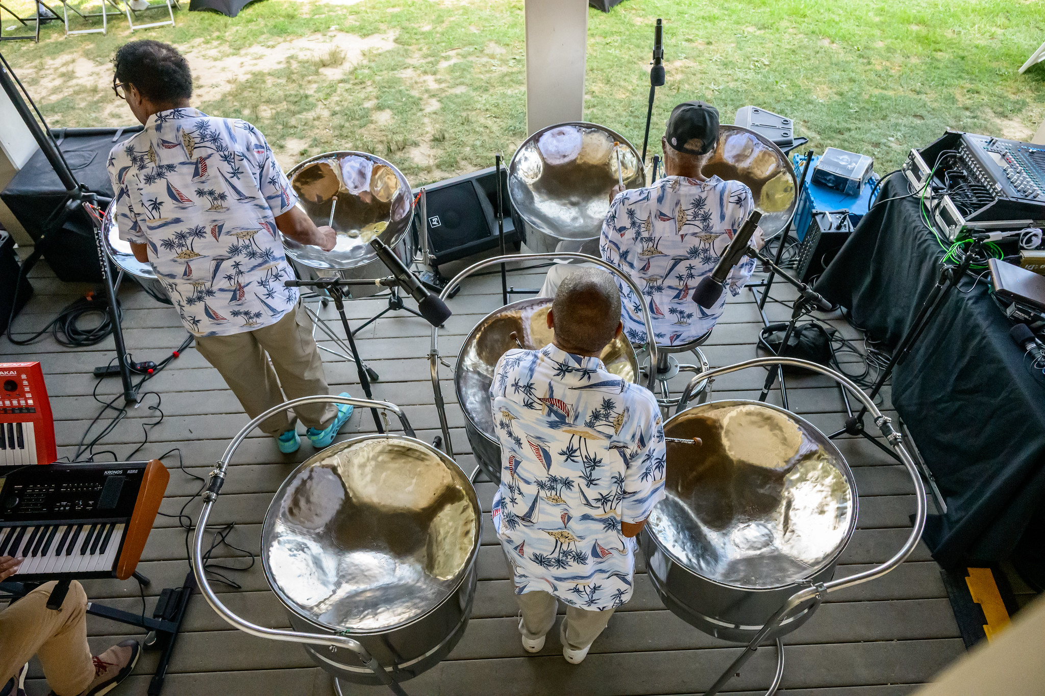 Three musicians wearing patterned shirts play steel drums on a wooden outdoor stage, surrounded by audio equipment, with grass visible in the background.