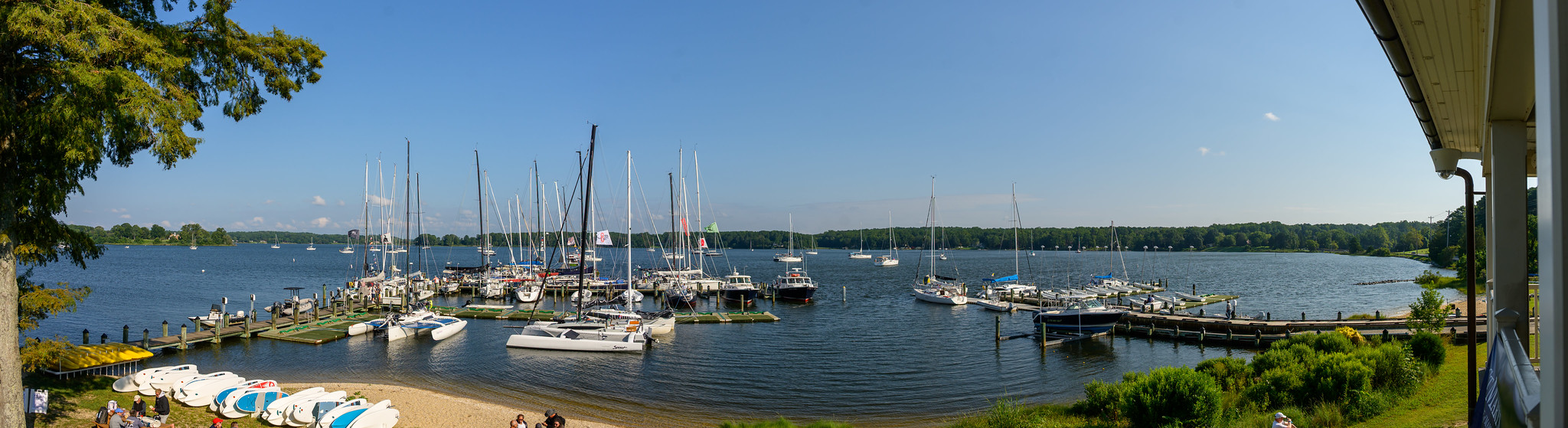Panoramic view of a marina with multiple sailboats docked, a small sandy beach with kayaks in the foreground, and calm water surrounded by greenery under a clear blue sky.