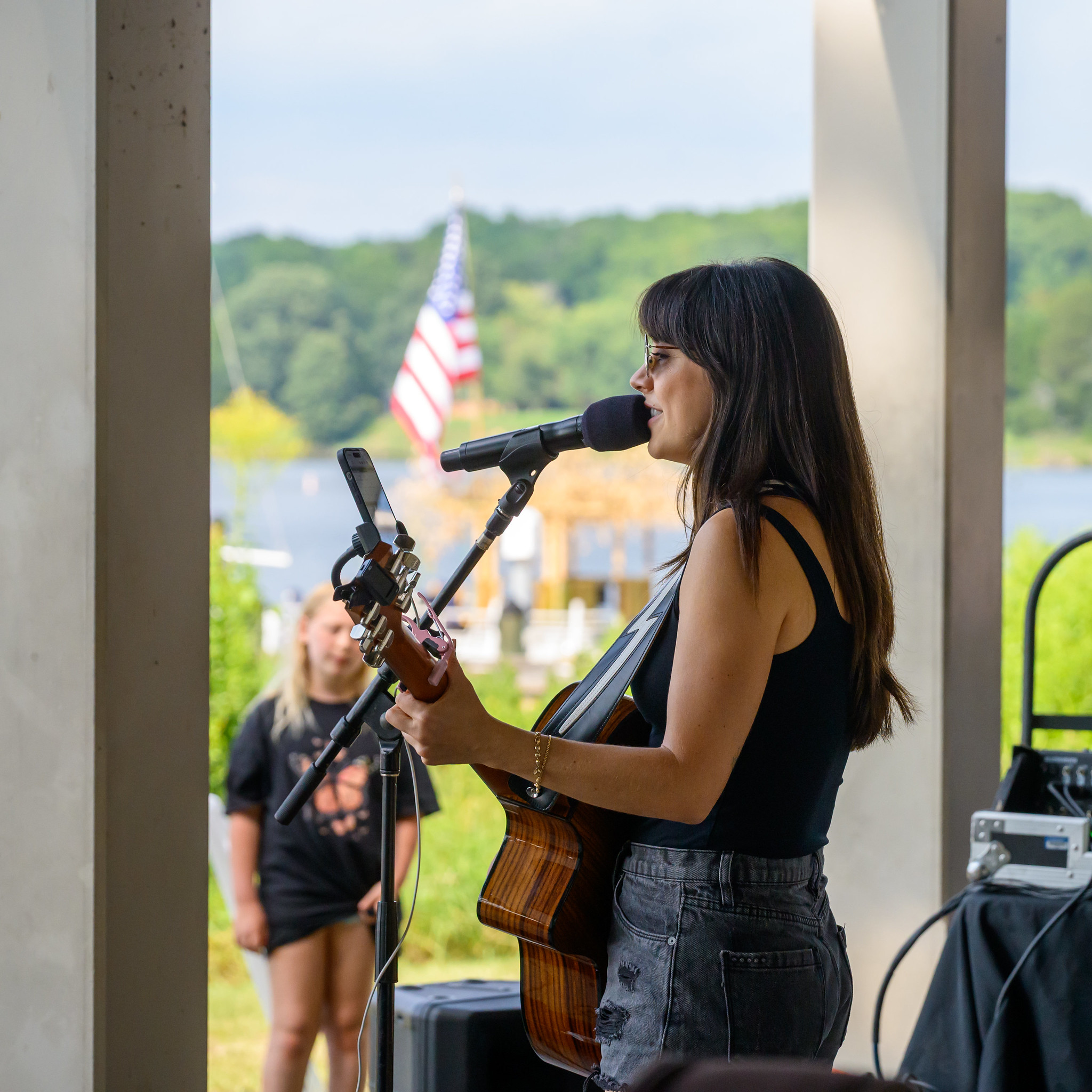 Woman playing acoustic guitar and singing into a microphone at an outdoor venue, with an American flag and greenery visible in the background.
