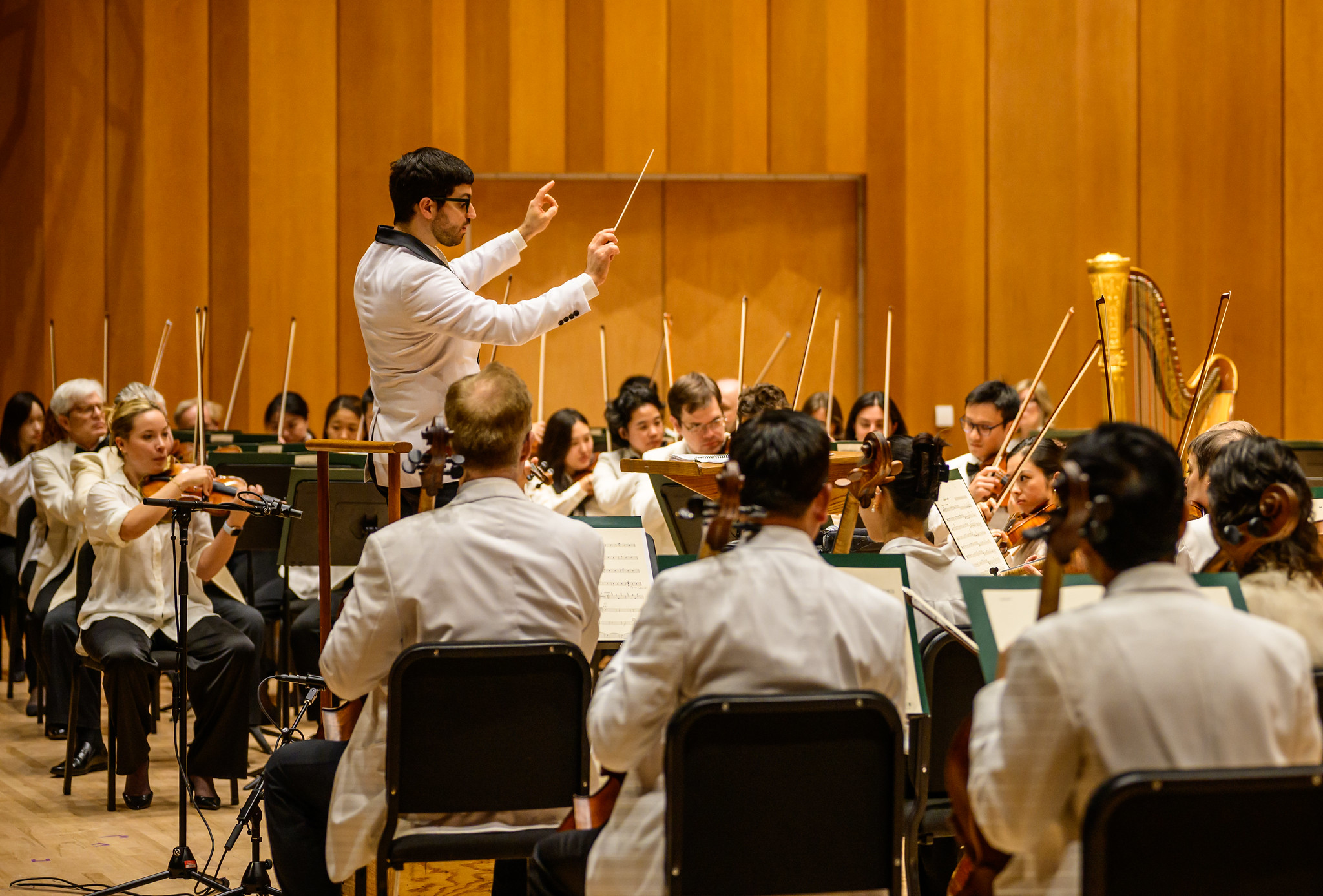 A conductor leads an orchestra of musicians dressed in white jackets, performing on stage with various instruments and a wooden background.