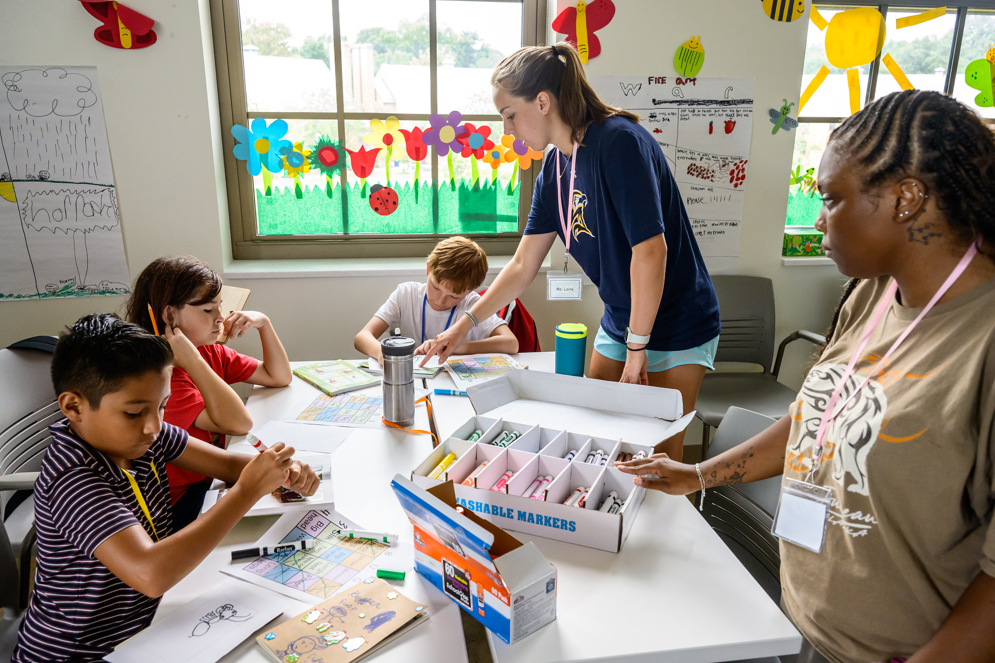 Three children sit at a table working on crafts with markers as two adults supervise in a bright classroom decorated with colorful art and paper bees.