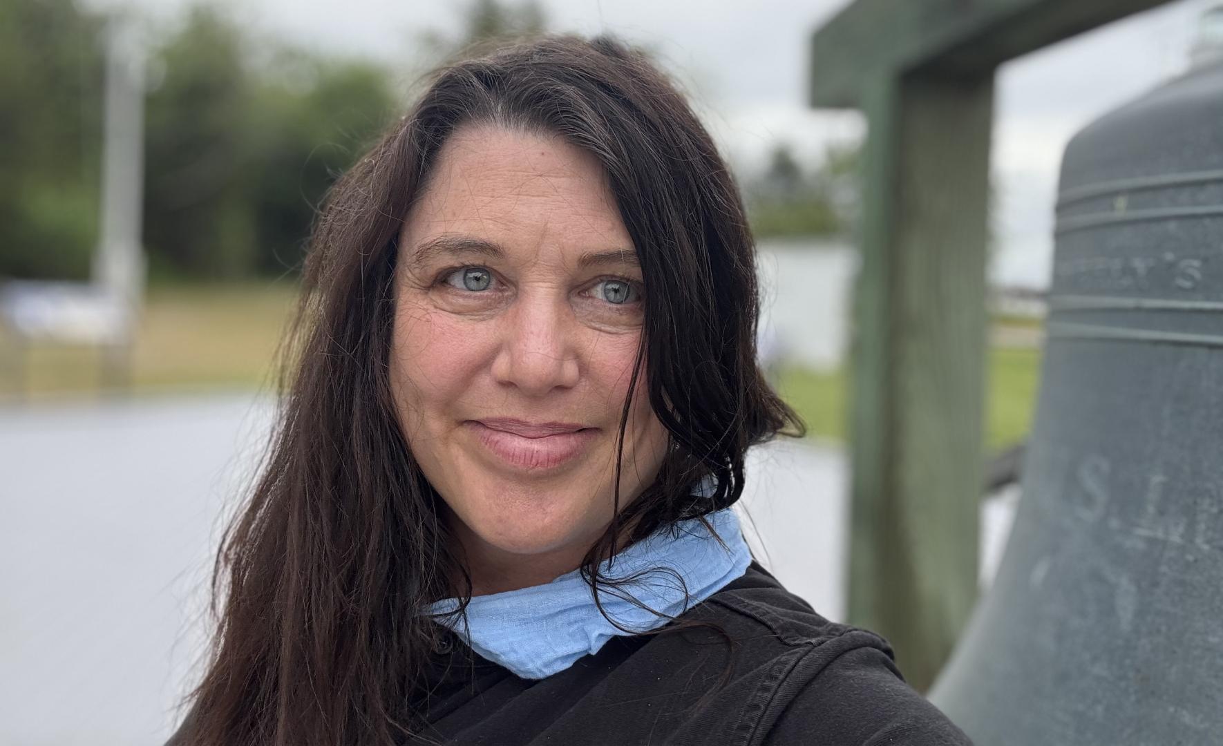 A woman with long dark hair stands outdoors near a large bell, looking at the camera and smiling slightly. She is wearing a black top and a blue scarf around her neck.