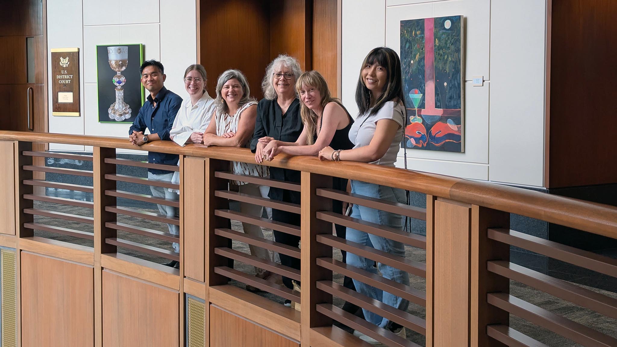 Six people stand in a row, leaning against a wooden railing inside a building with art displayed on the walls behind them.