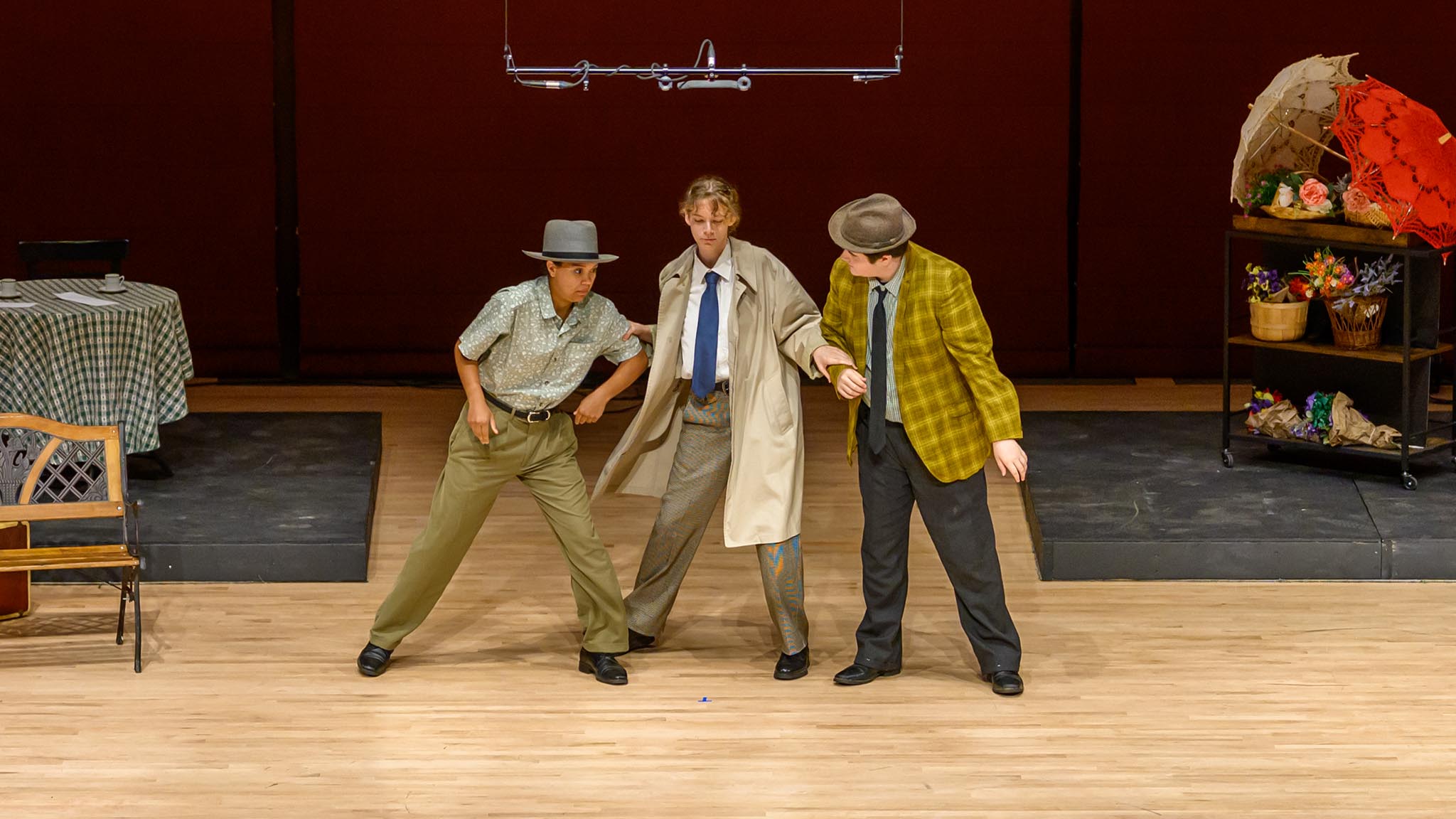 Three actors on stage perform a dramatic scene; one wears a trench coat, the others wear hats and patterned jackets. Stage props and furniture are visible in the background.