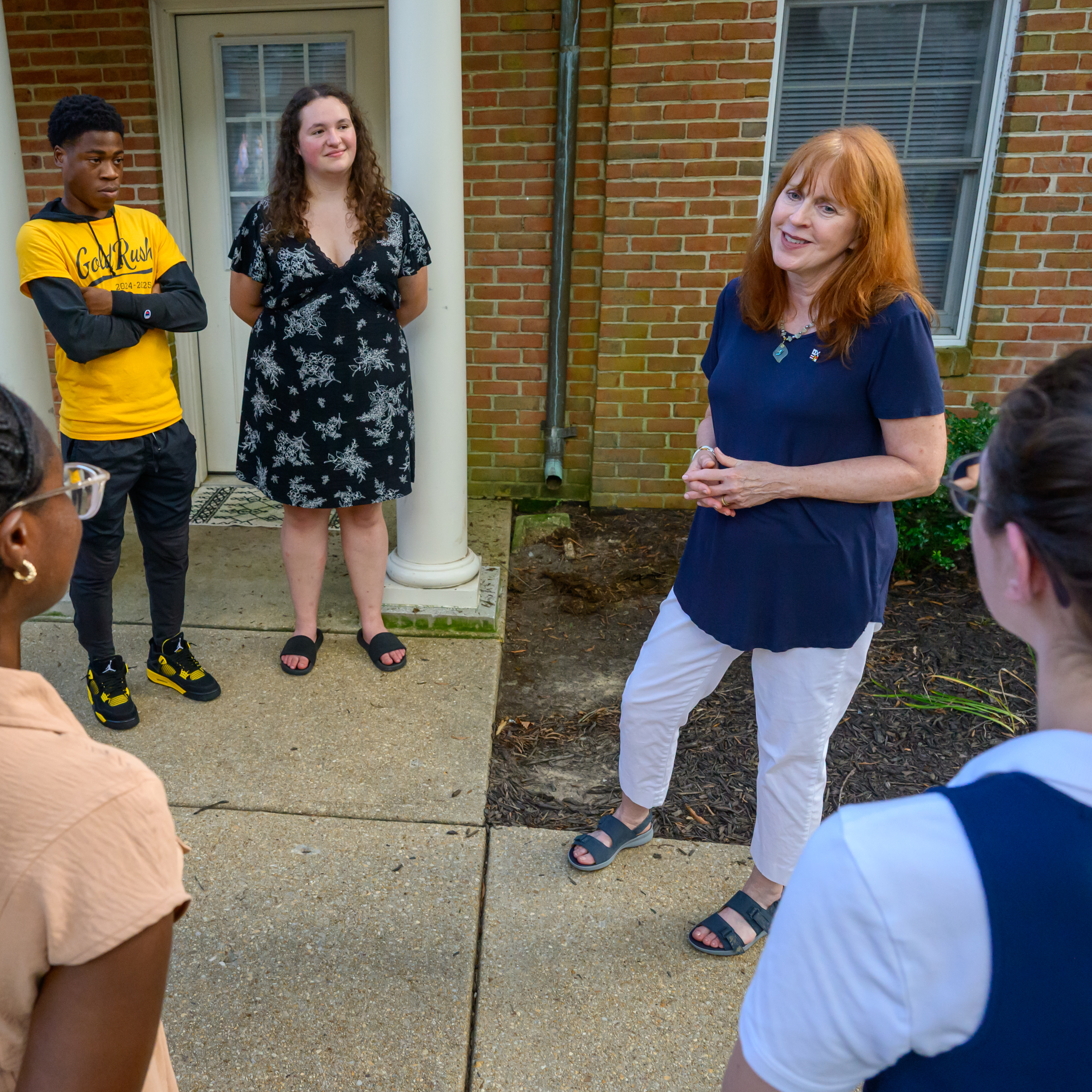 A woman speaks to a small group of people standing in a semicircle outside a brick building with a white pillar and window.