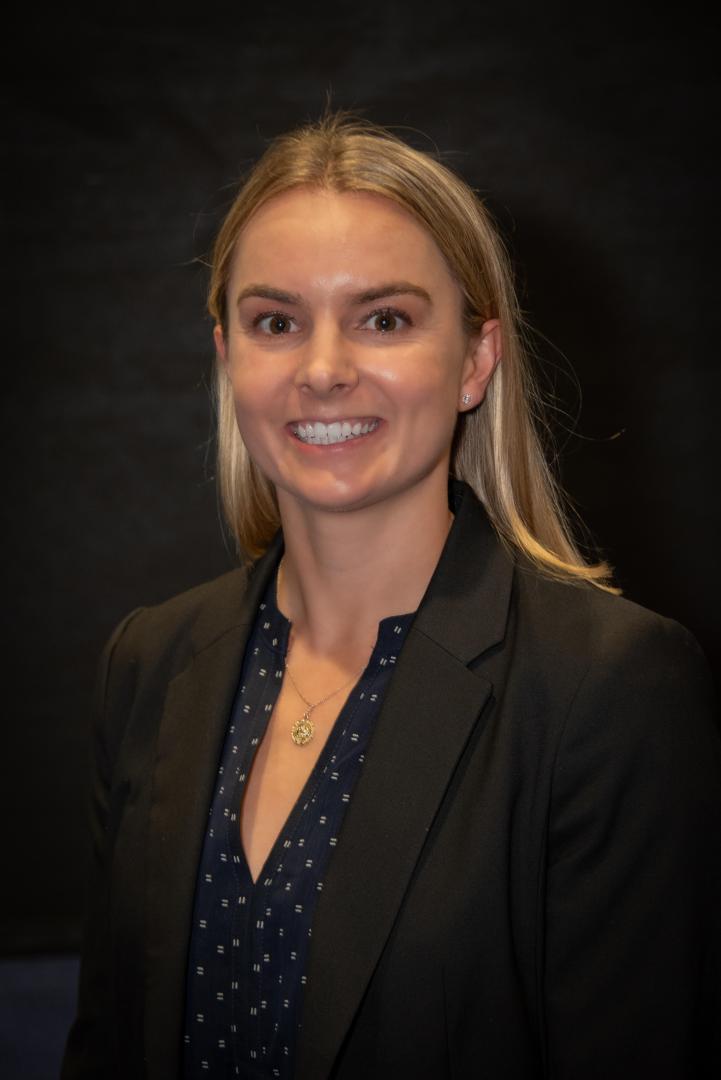 Claire Robey, a woman with straight blonde hair, wearing a dark blazer over a patterned blouse, smiles in front of a plain dark background.