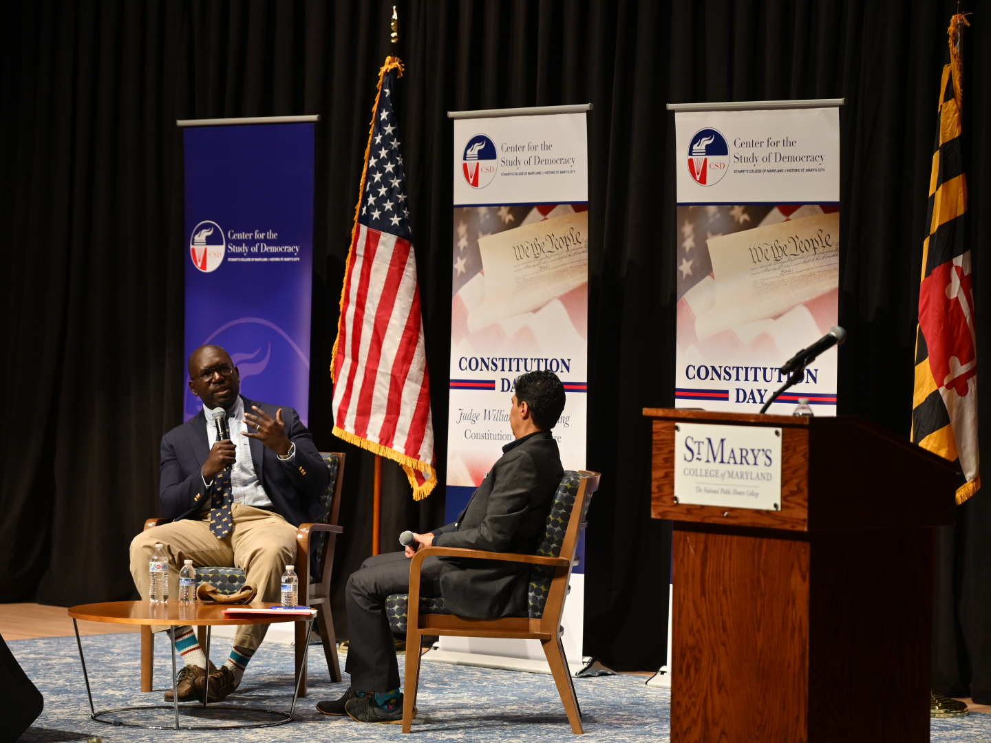 Two people sit on stage having a discussion during a Constitution Day event at St. Mary's College of Maryland, with banners and an American flag in the background.