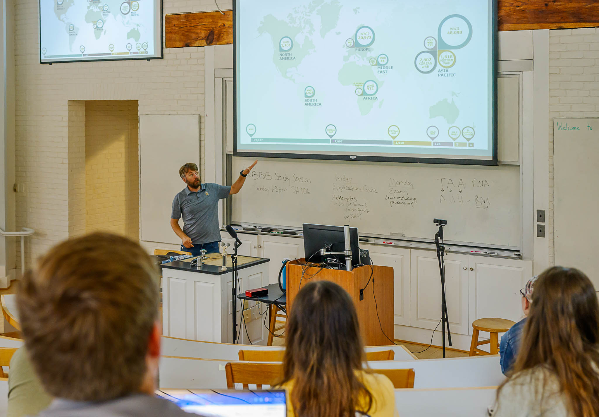 A man stands at the front of a classroom, pointing at a world map on a projector screen, while students sit facing him and take notes.