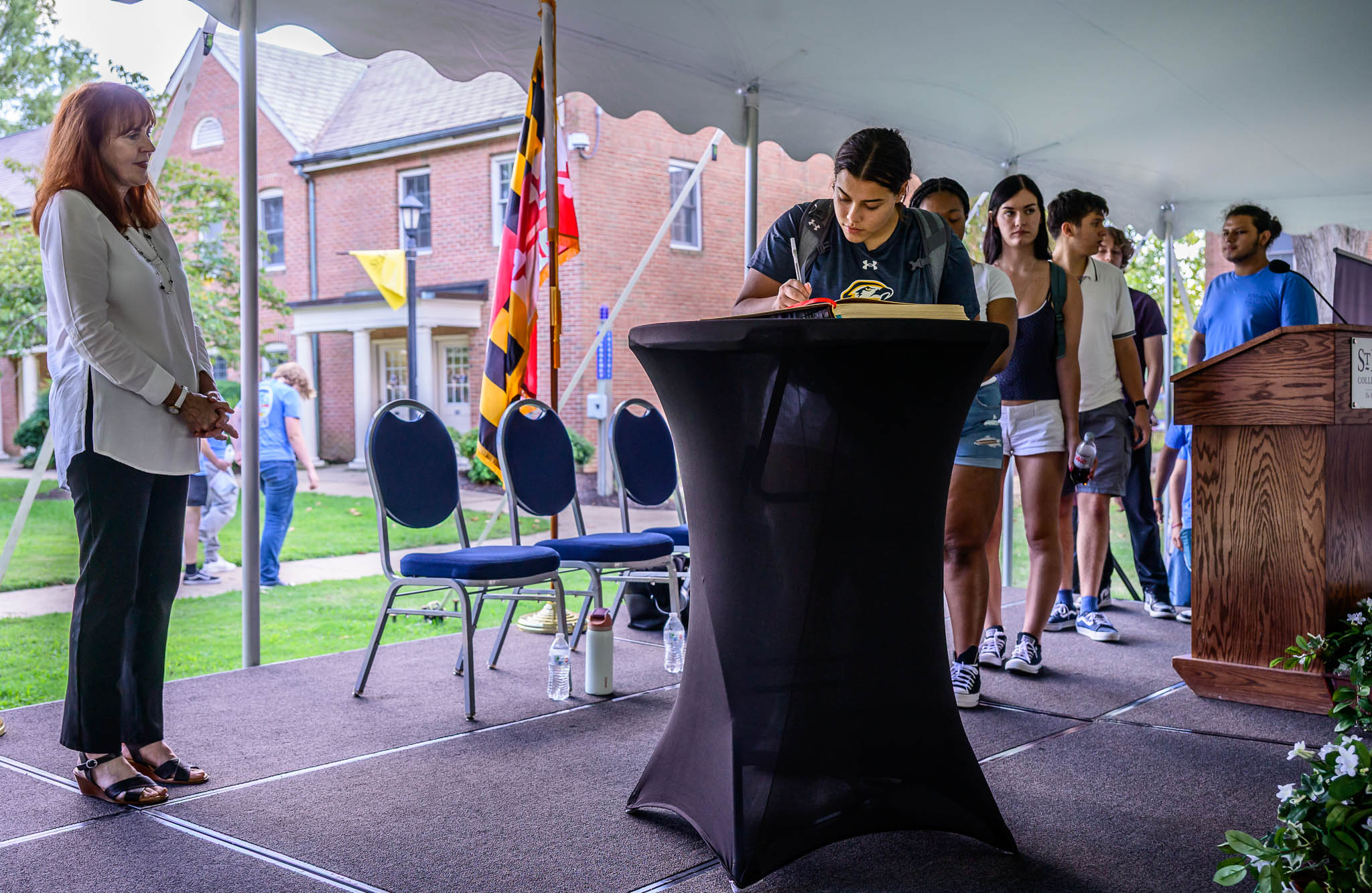 A student signs a book on a podium while others wait in line; an adult stands to the side on a stage set up outdoors with chairs and flags visible.